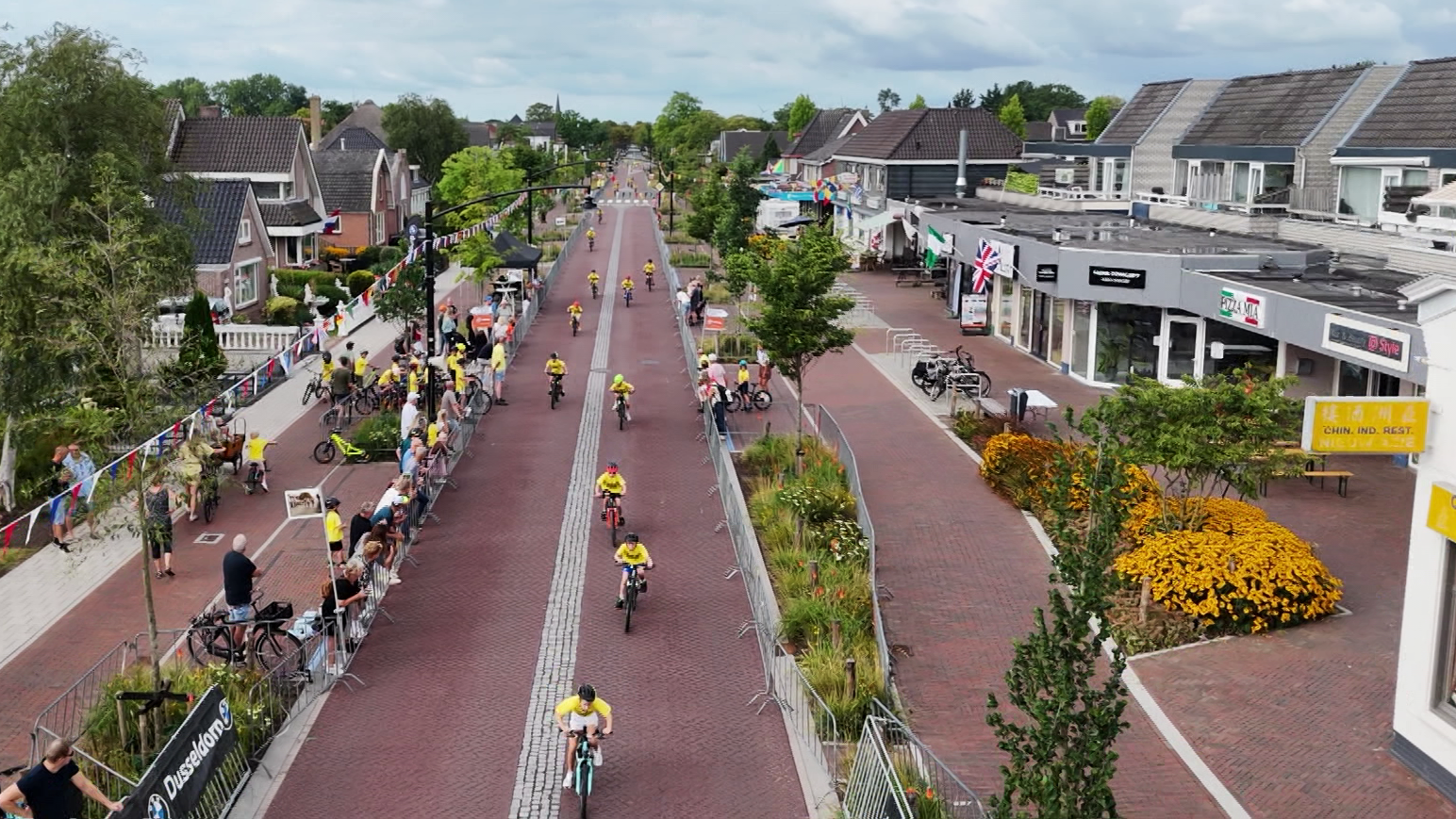 Wielerwedstrijd in dorpsstraat, renners in gele shirts fietsen langs afzettingen en toeschouwers, met winkels rechts
