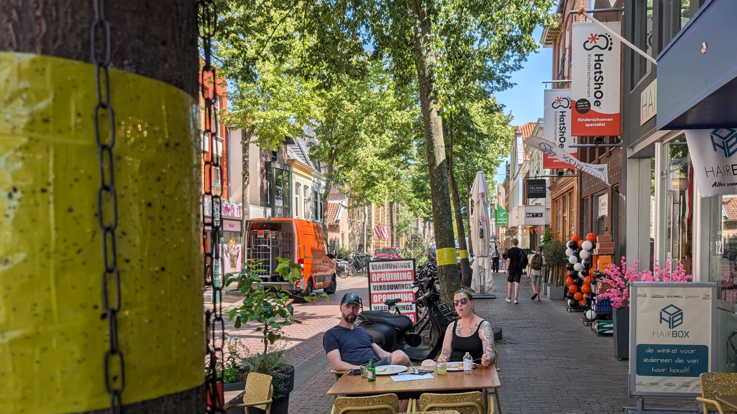 Twee mensen zitten aan een terrastafel in een winkelstraat met bomen, fietsen en winkelborden, waaronder HatShoe