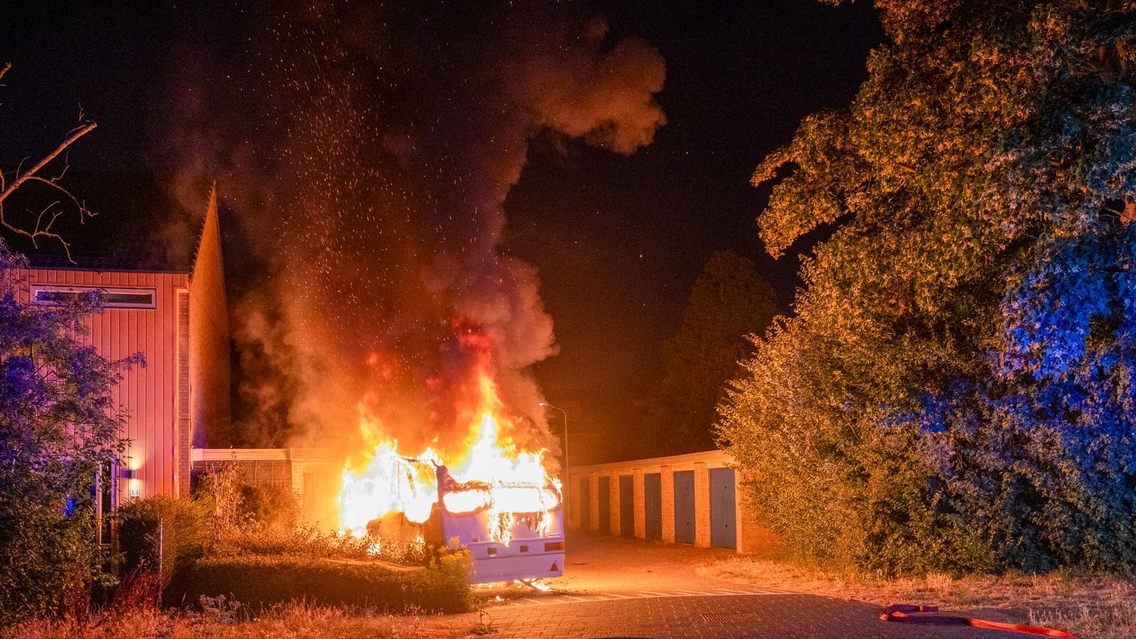 Brandende camper naast een gebouw in de nacht, met hoge vlammen en dikke rookpluim langs een rij garageboxen