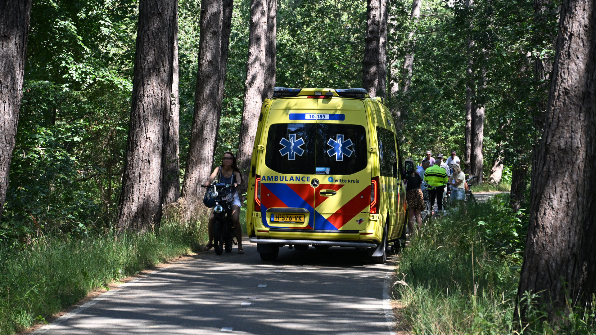 Gele ambulance op een bospad, met fietsers en omstanders eromheen tussen hoge bomen