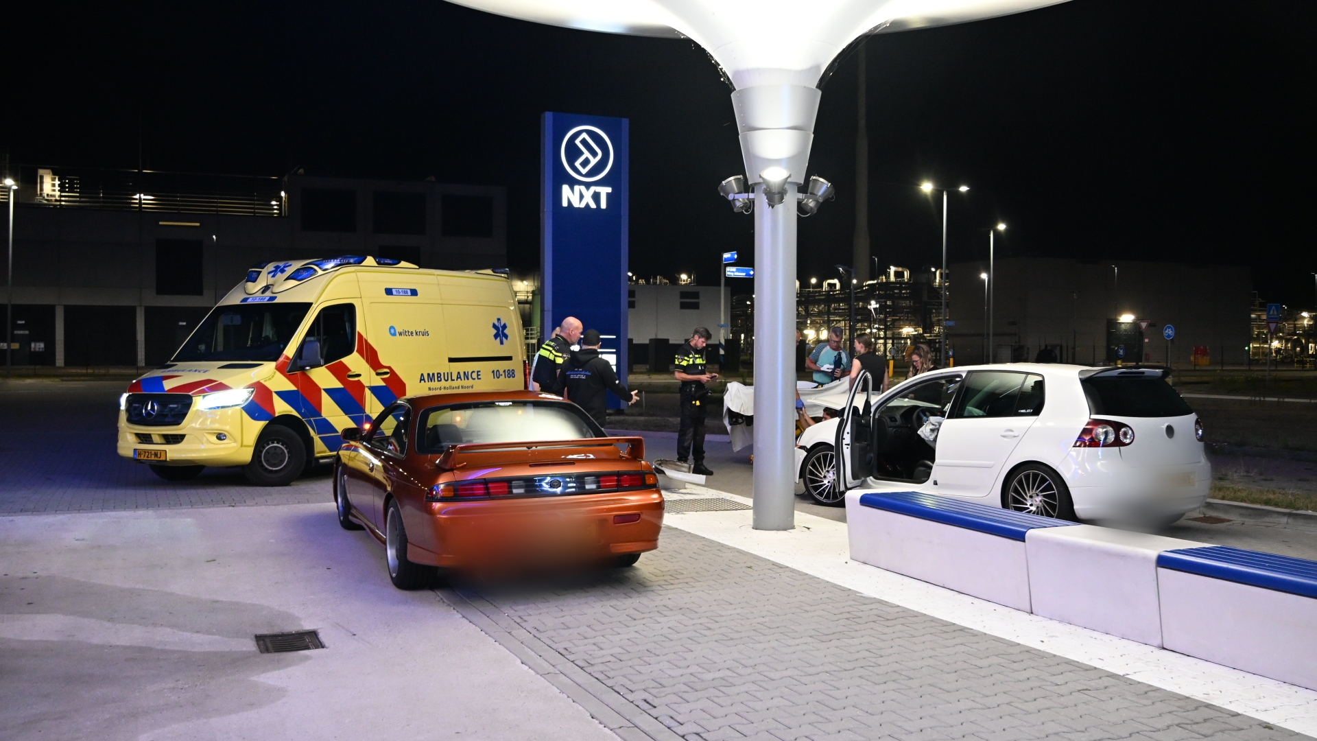 Ambulance en politie bij twee auto’s na een incident bij NXT, met een witte auto met open portier en een rode coupé