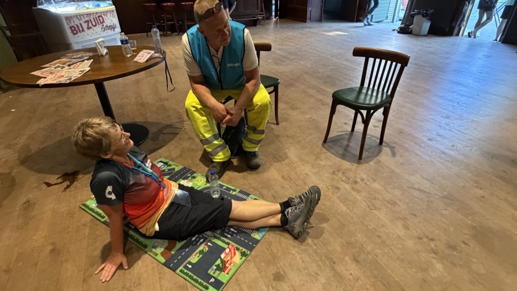 Man rust uit op mat op de vloer terwijl een vrijwilliger in veiligheidspak naast hem zit in een zaal met tafels en stoelen