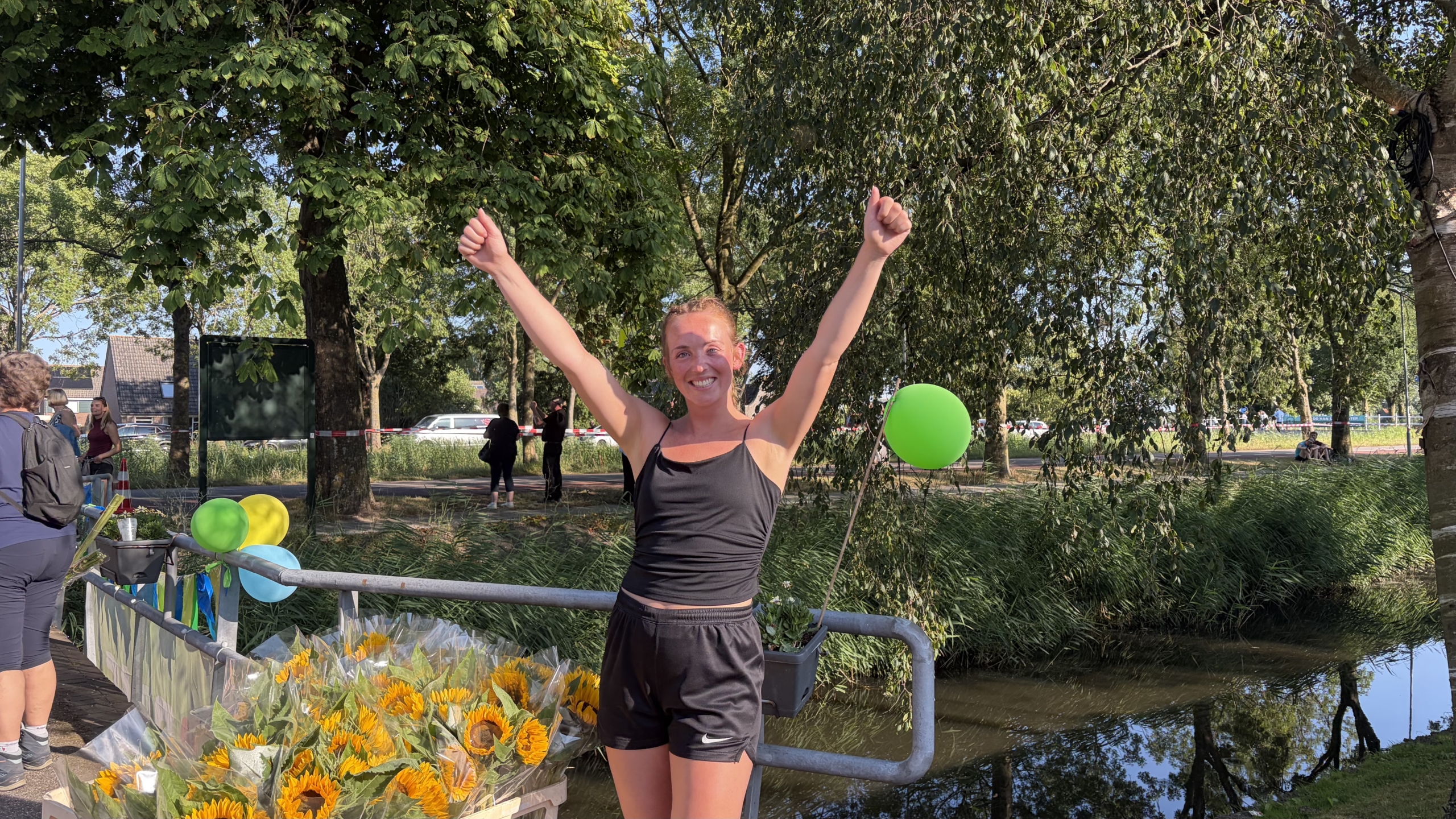 Vrolijke hardloopster met beide armen omhoog bij de finish, naast zonnebloemen en groene ballonnen langs een sloot