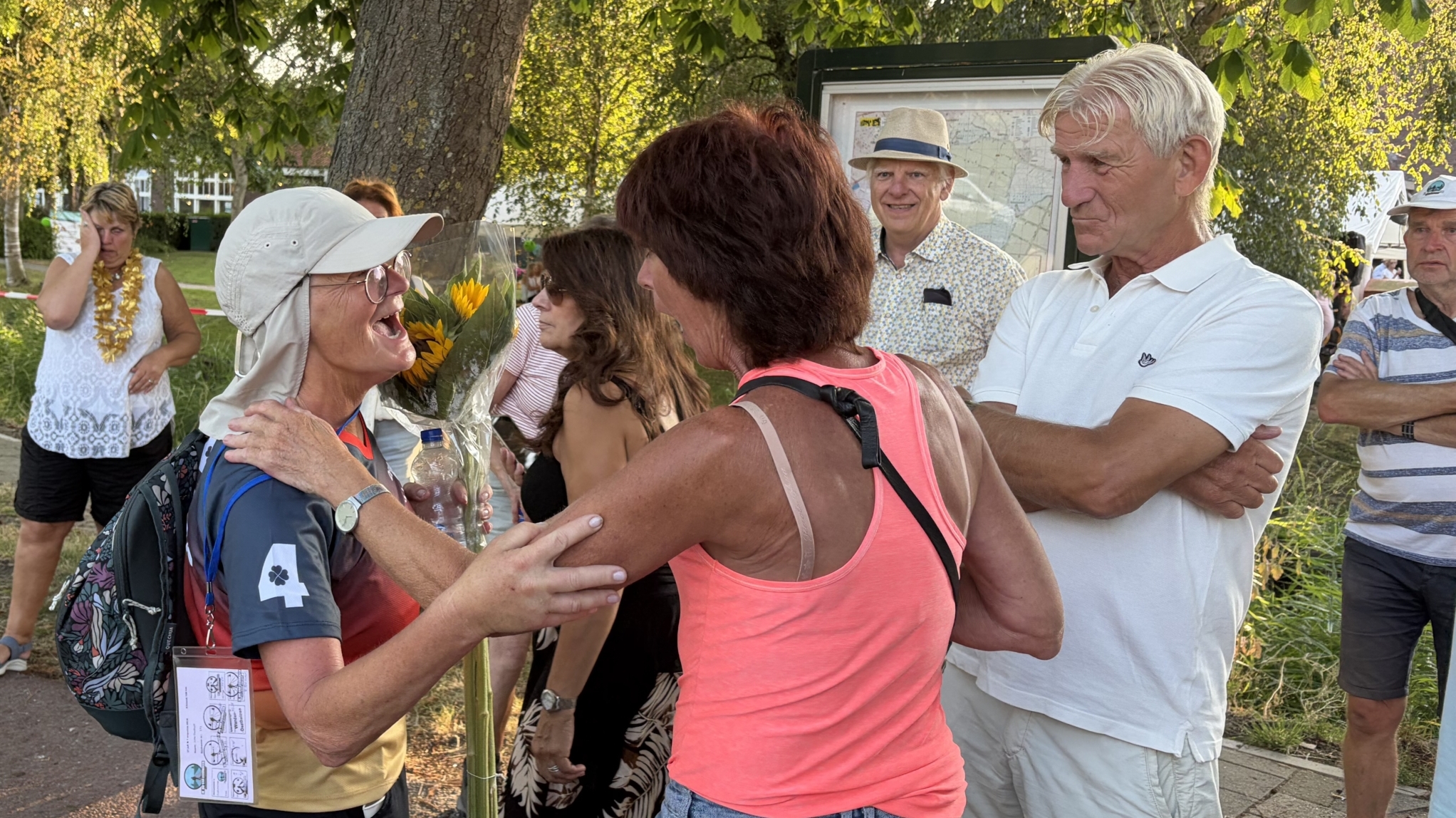 Vrouw met pet en rugzak krijgt een bos zonnebloemen en een omhelzing, omringd door toeschouwers langs de weg