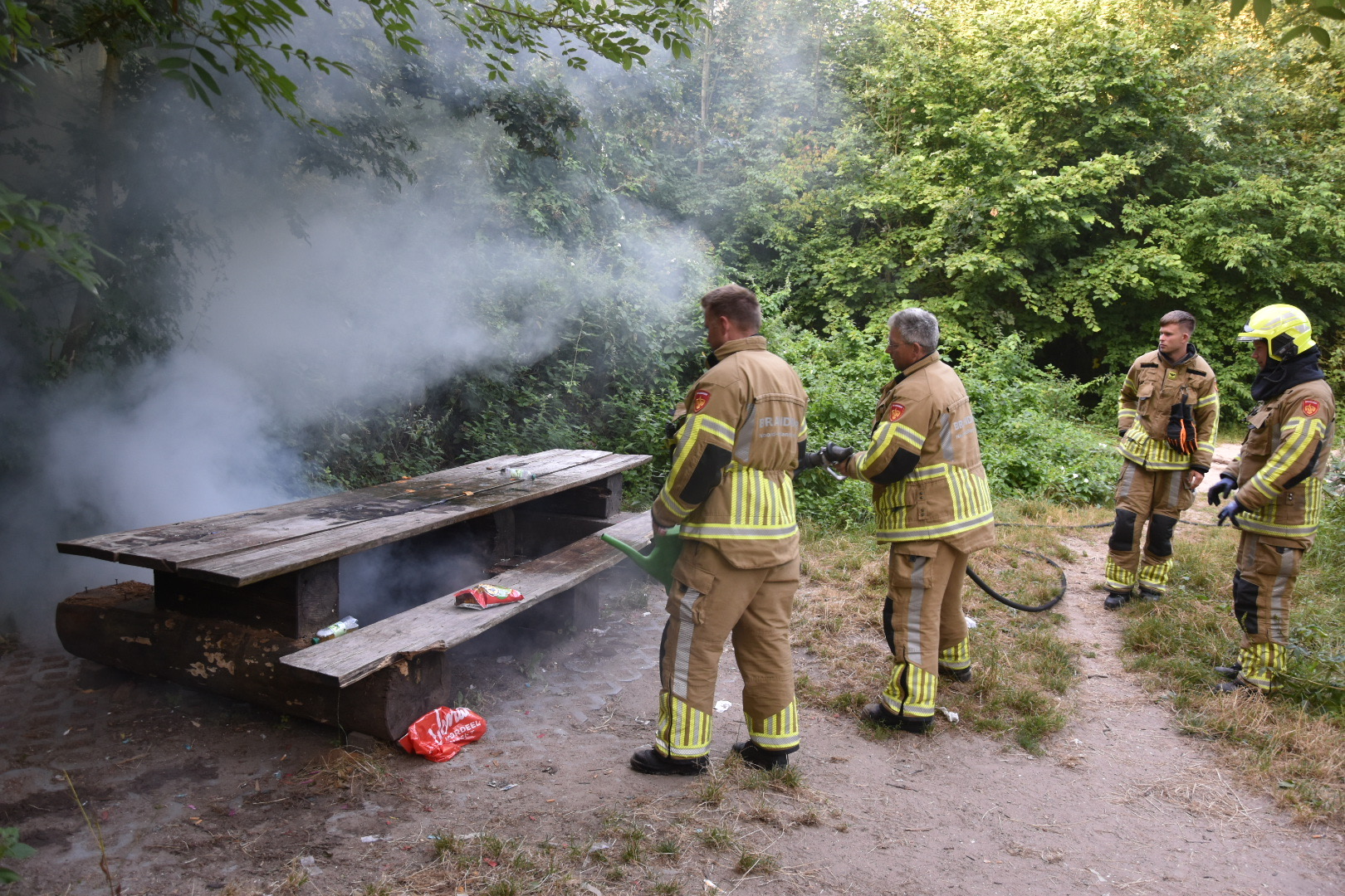 Brandweerlieden blussen een smeulende picknicktafel in het bos terwijl rook tussen de bomen hangt