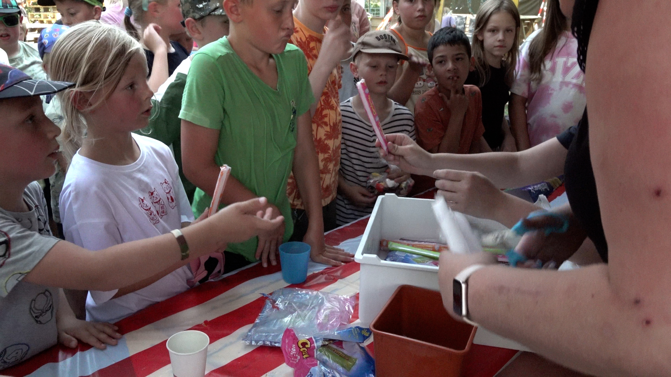 Groep kinderen staat rond een tafel terwijl een volwassene snoep uitdeelt uit een bak