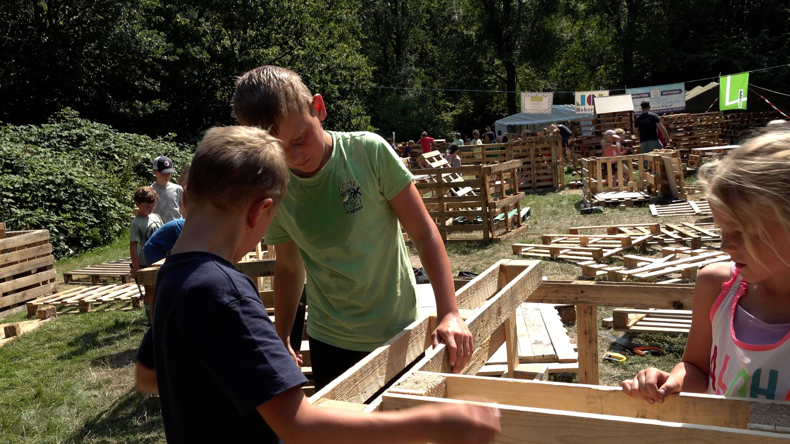 Kinderen bouwen met houten pallets op een zonnig buitenterrein, met meer palletconstructies en mensen op de achtergrond