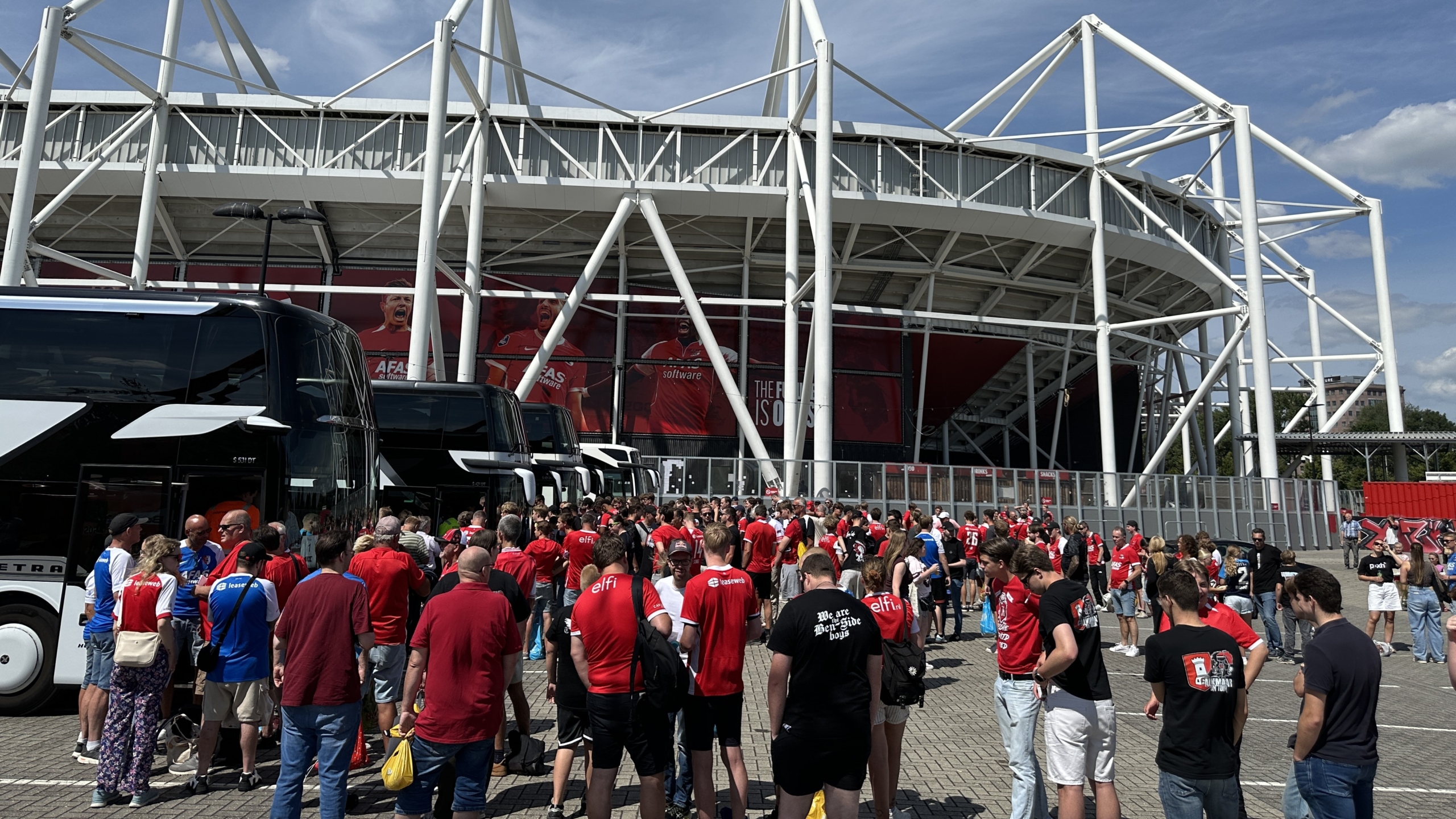 Drukke menigte voetbalsupporters bij een stadion met witte staalconstructie en geparkeerde touringcars
