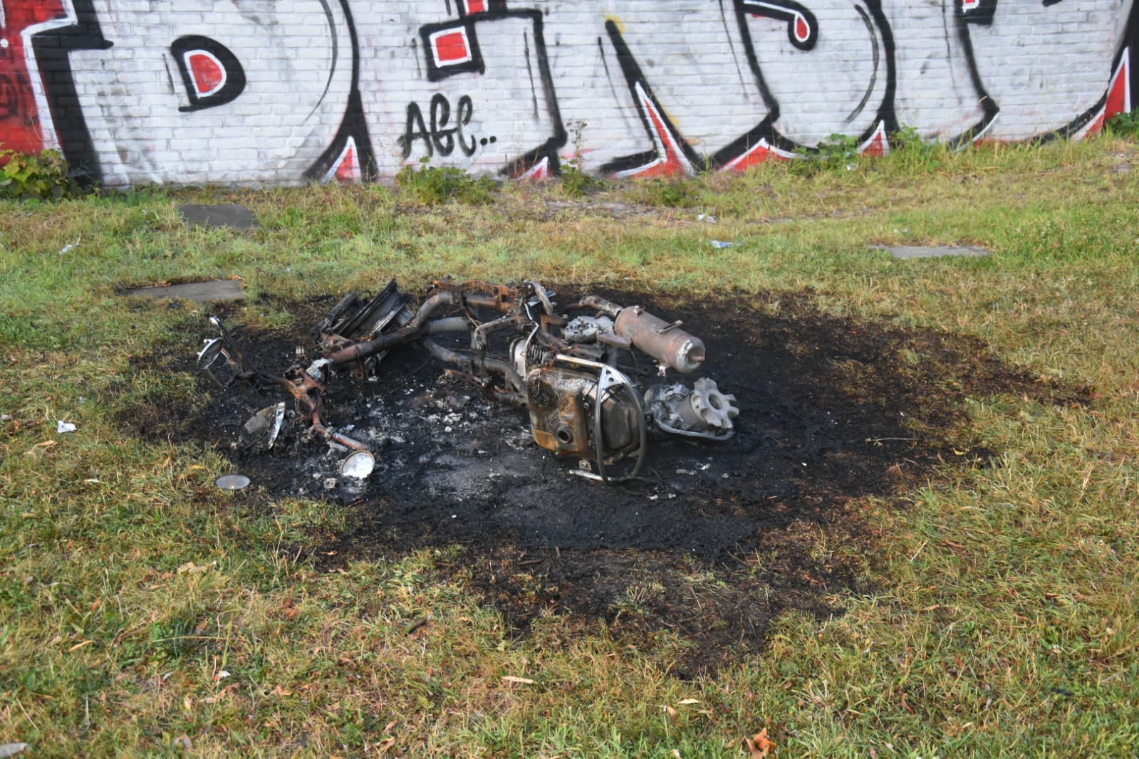 Uitgebrande motor- of brommerresten op een zwartgeblakerde plek in het gras voor een muur met graffiti