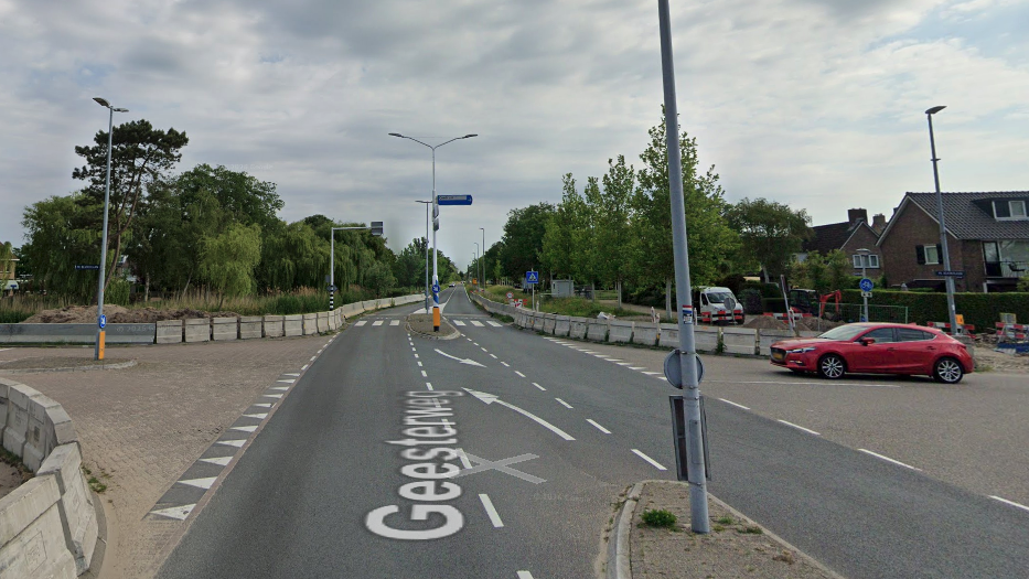 Kruispunt op een brede weg met wegwerkzaamheden en afzettingen; rode auto rijdt langs huizen onder bewolkte lucht