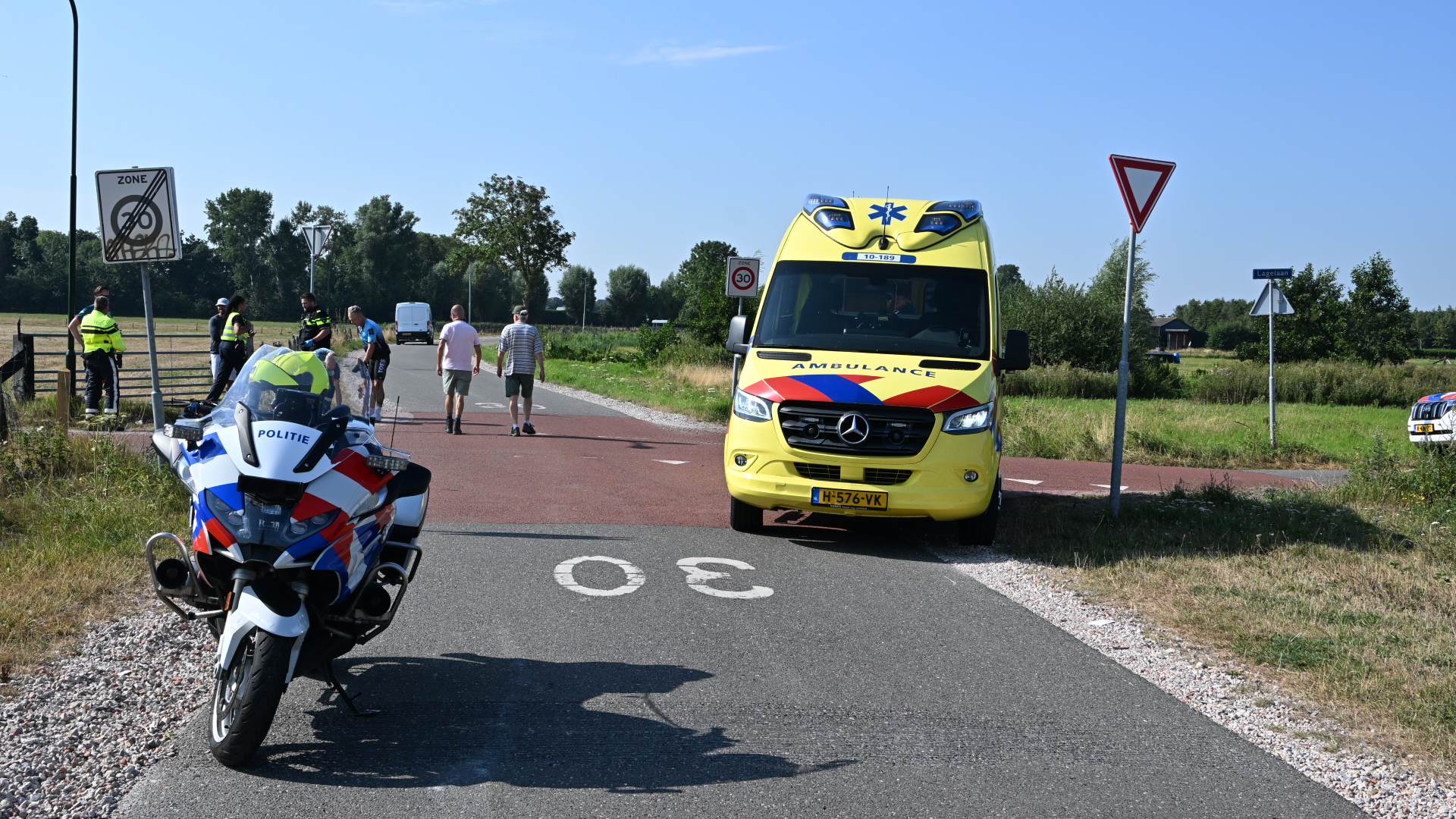 Politiemotor en ambulance staan op een landelijke weg bij een incident, met hulpverleners en omstanders op de achtergrond