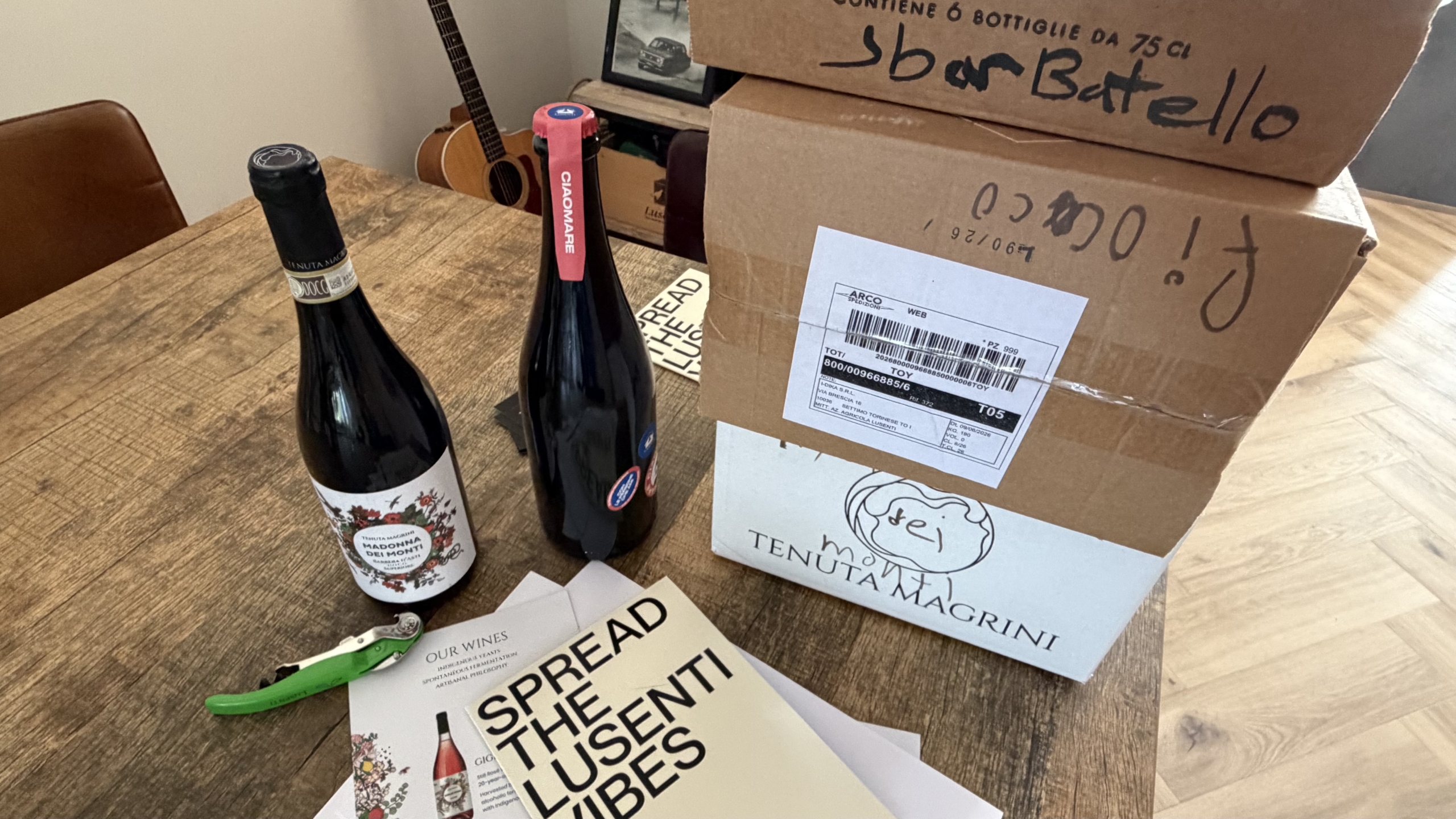 Twee wijnflessen naast gestapelde verzenddozen op een houten tafel, met kurkentrekker en brochure “SPREAD THE LUSCIOUS VIBES”
