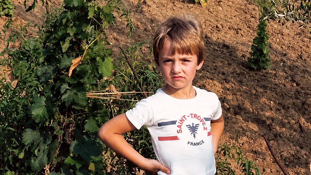 Jongen met handen in de zij in een moestuin, met druivenrank en omgewoelde aarde op de achtergrond