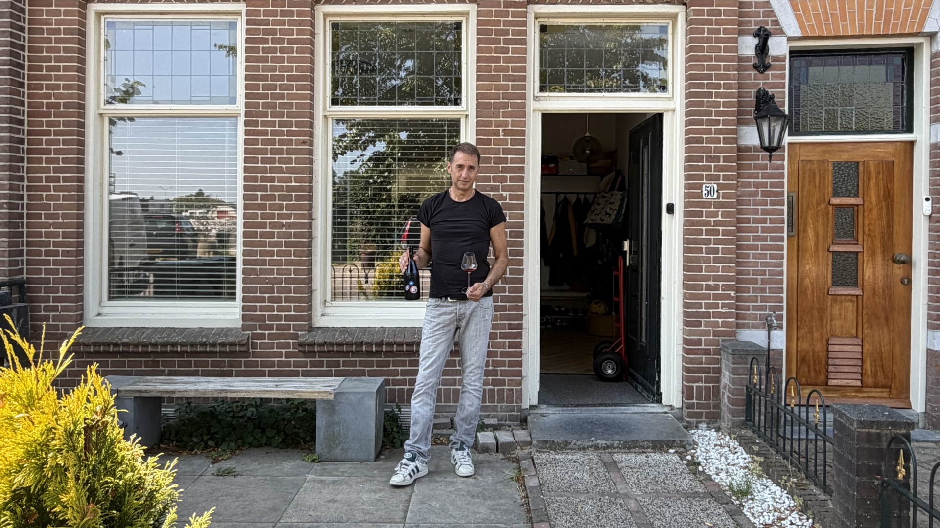 Man staat voor een bakstenen huis met open voordeur en houdt een fles en wijnglas vast naast huisnummer 50