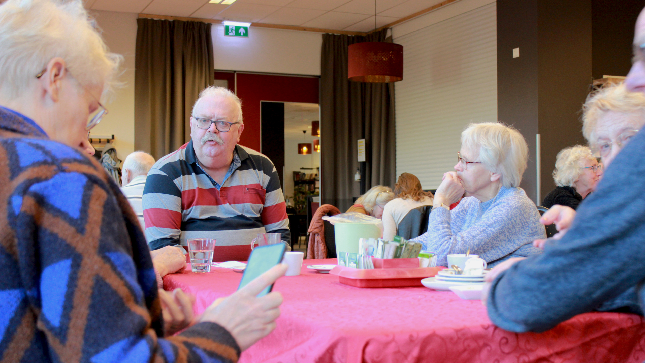 Groep ouderen zit aan een tafel met rood tafelkleed en koffiekopjes in een ontmoetingsruimte, in gesprek