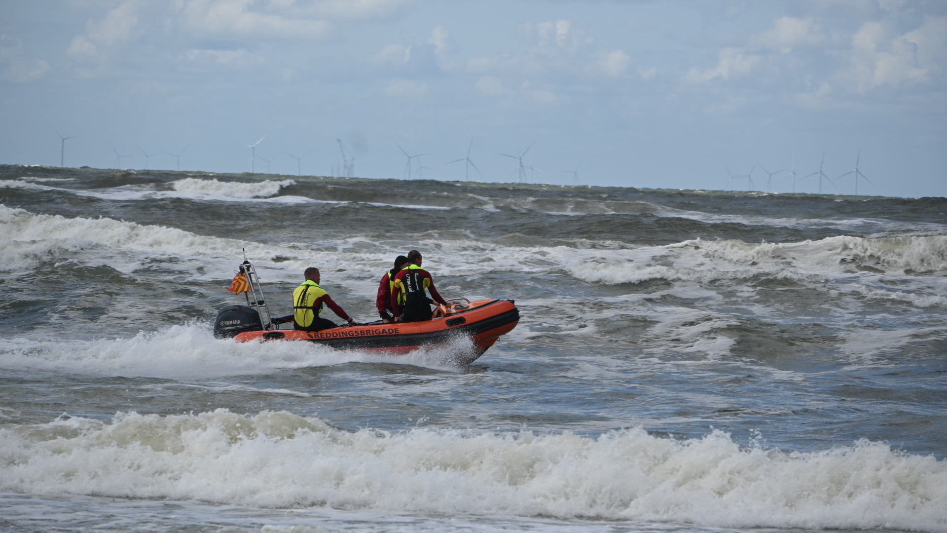 Reddingsbrigade in oranje rubberboot vaart over ruwe zee, met windturbines op de horizon