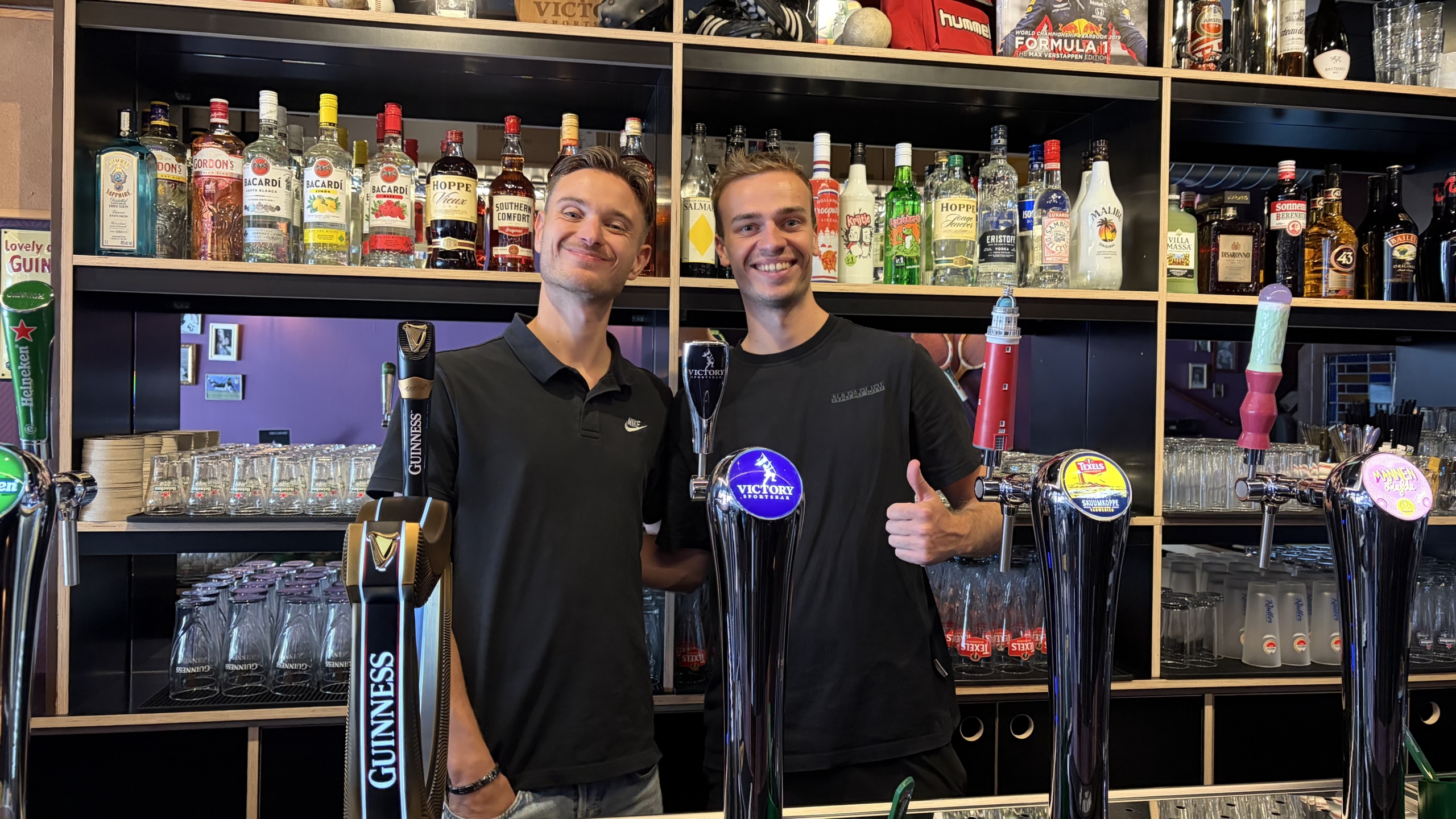 Twee barmedewerkers glimlachen achter de bar met bierpompen, met flessen sterke drank en glazen op de achtergrond