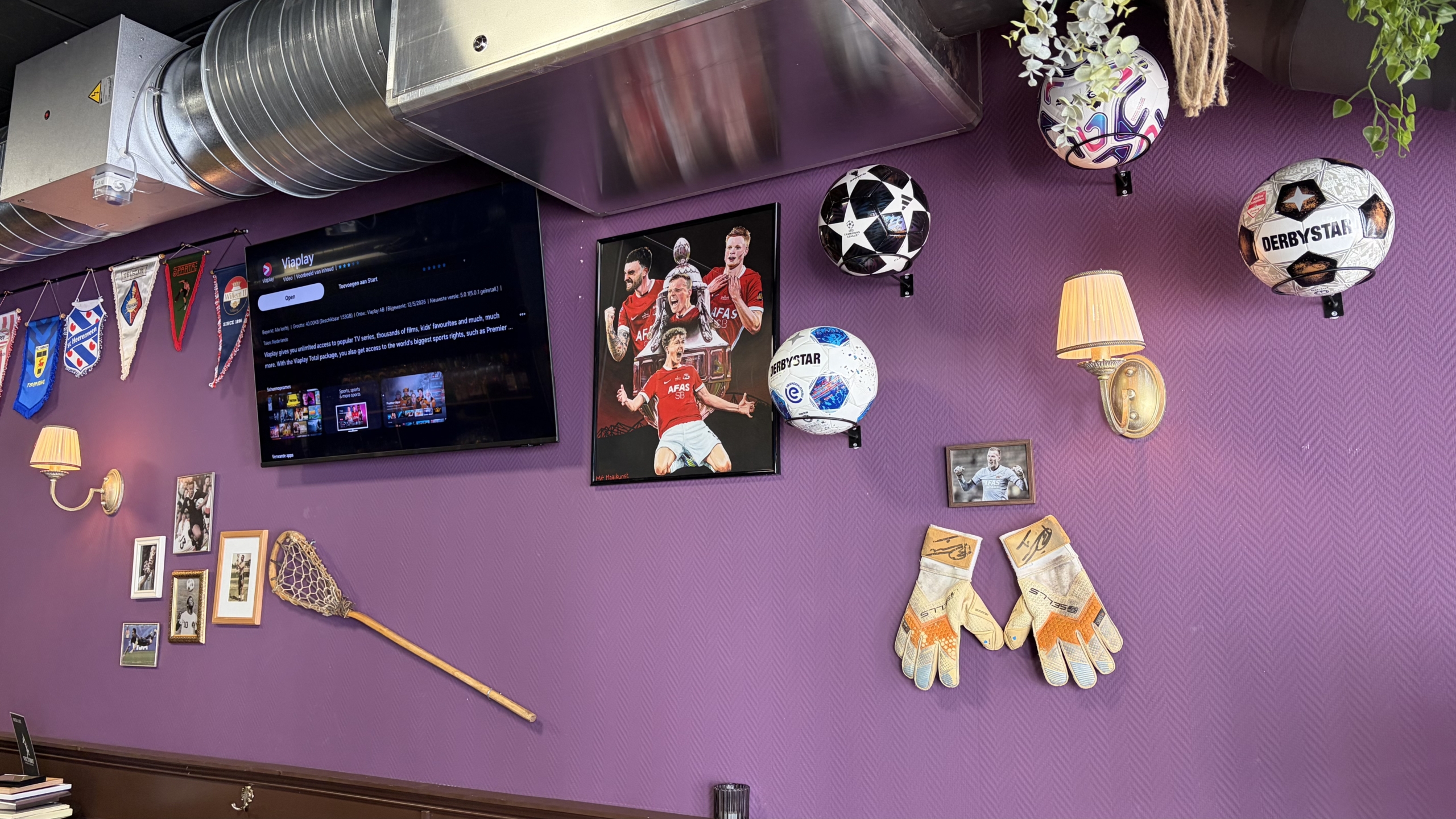 Paarse muur met sportdecoratie: tv-scherm, voetbalposters, meerdere voetballen, vaantjes, foto’s en keepershandschoenen