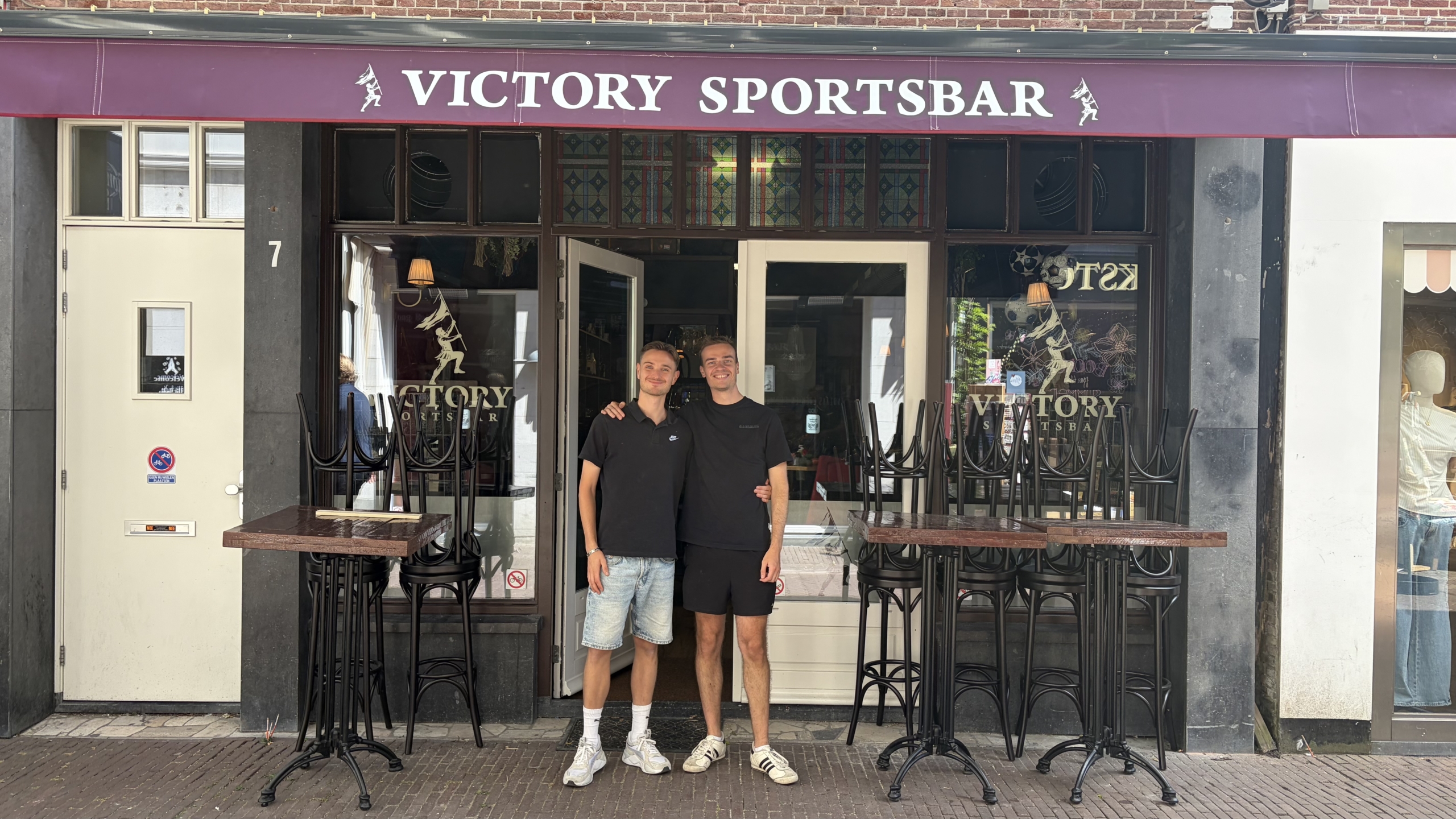 Twee mannen poseren arm in arm voor de ingang van Victory Sportsbar, met gestapelde barkrukken en tafels buiten