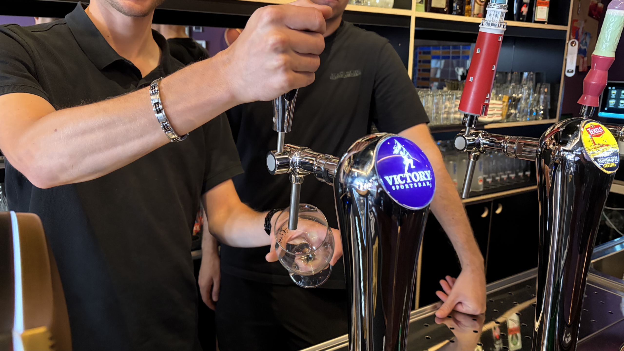 Barman tapt bier in een glas aan de bar, met glanzende tapkranen en het logo “Victory Sportsbar” zichtbaar