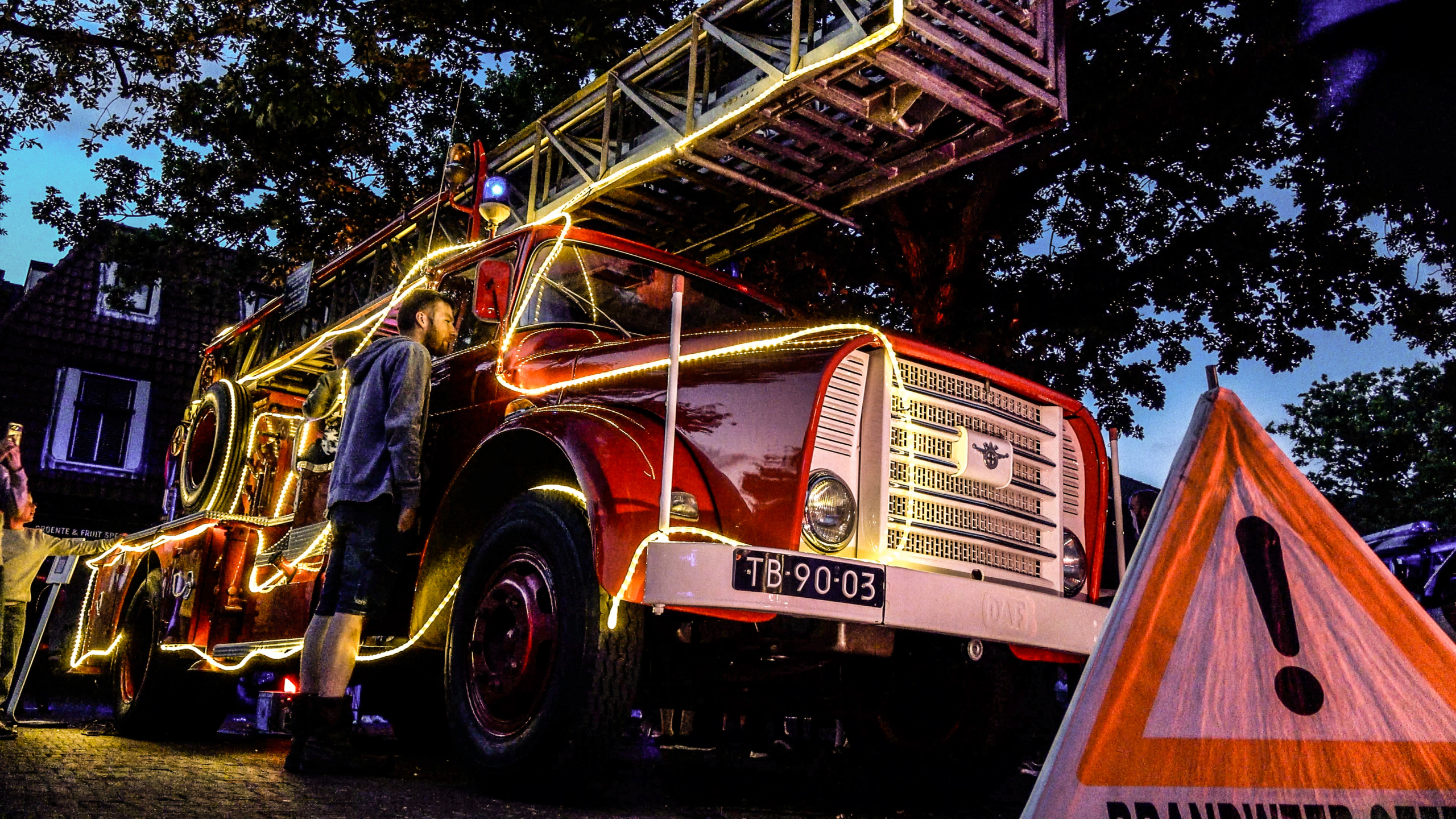 Rode brandweerwagen met uitgeschoven ladder, versierd met lichtsnoeren, terwijl mensen er ’s avonds naast staan