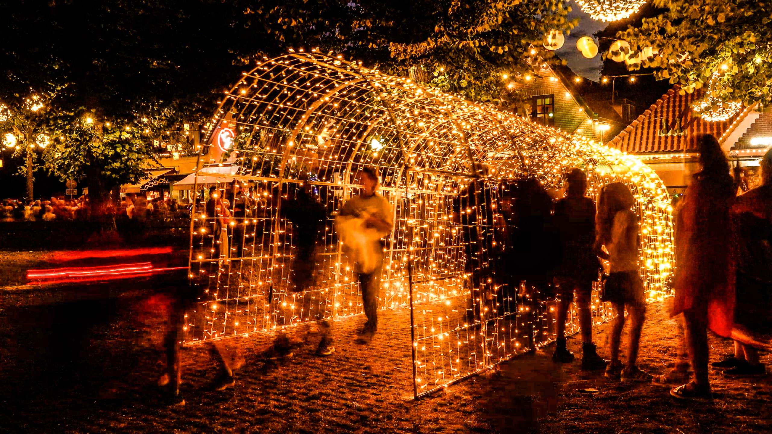 Verlichte lichttunnel van warmwitte kerstlampjes in een park bij nacht, met voorbijlopende mensen en sfeerverlichting in bomen