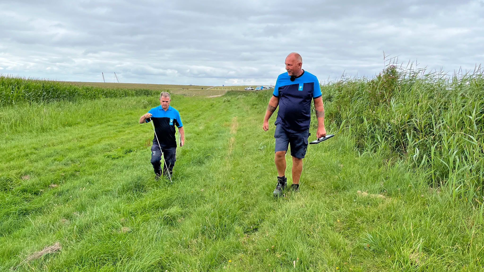Twee mannen in blauwe werkkleding lopen over een grasdijk, één met meetlint en één met een apparaat in de hand
