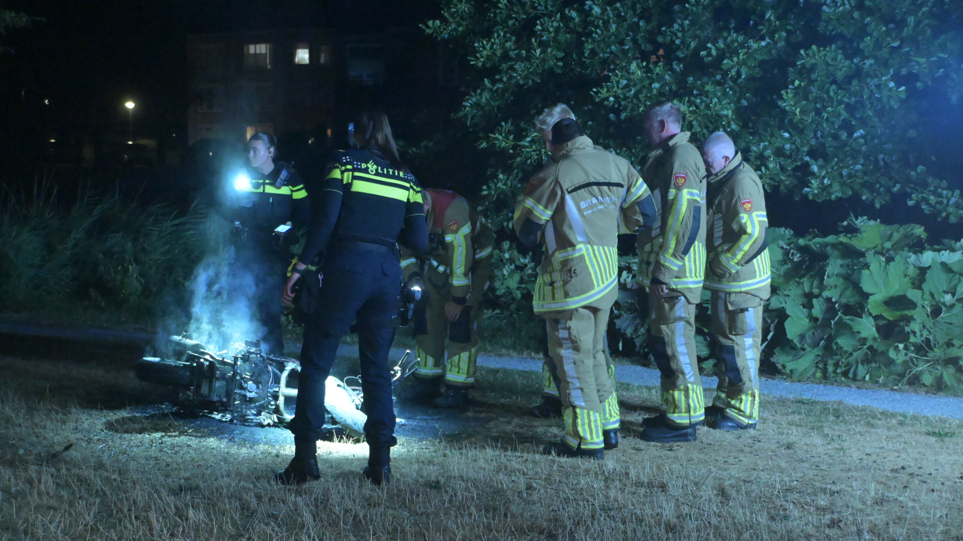 Politie en brandweer onderzoeken ’s nachts een rokende motorfiets die op zijn kant ligt in het gras