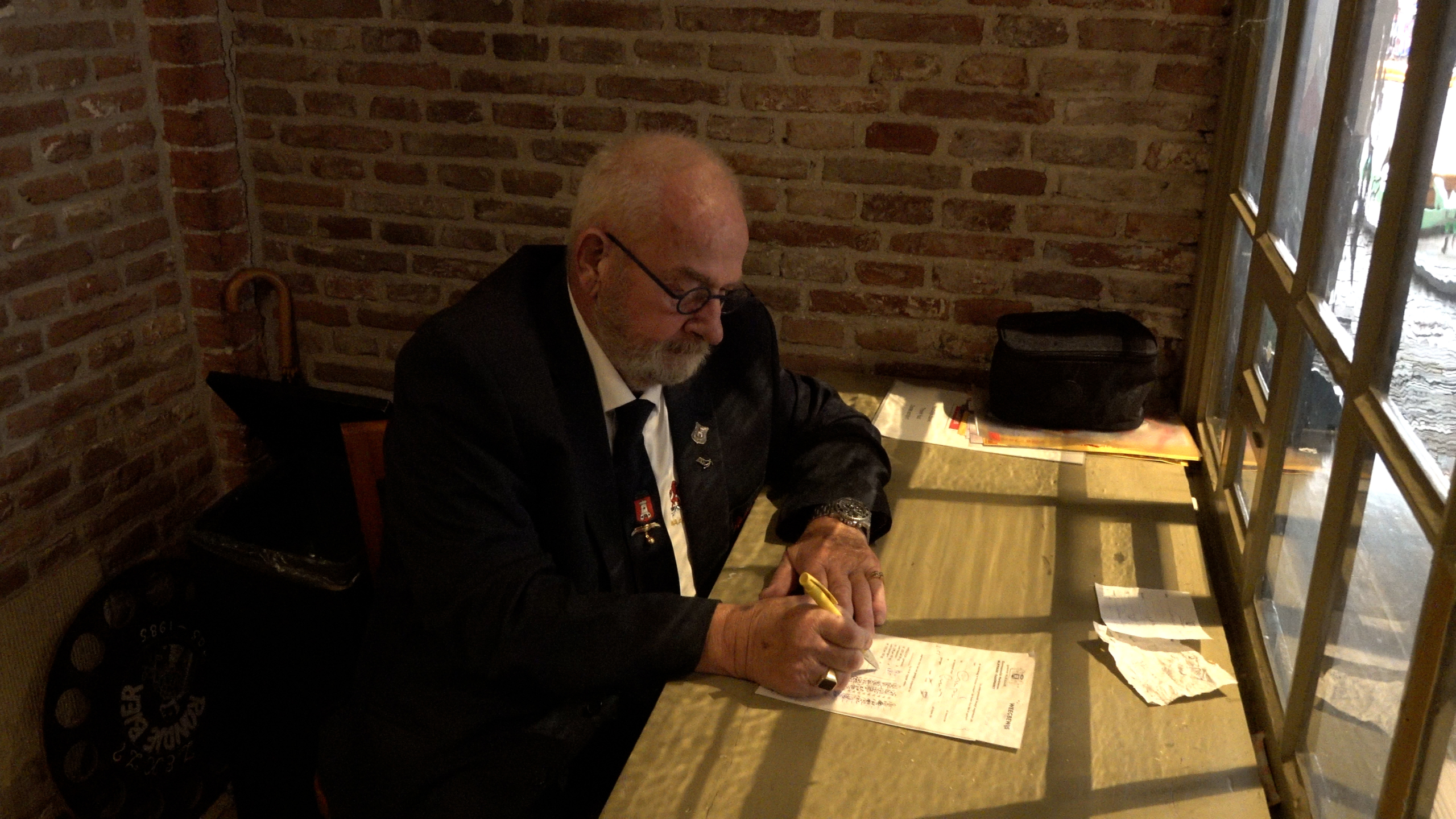 Oudere man in pak schrijft met een pen op papier aan een tafel bij het raam, met bakstenen muur op de achtergrond