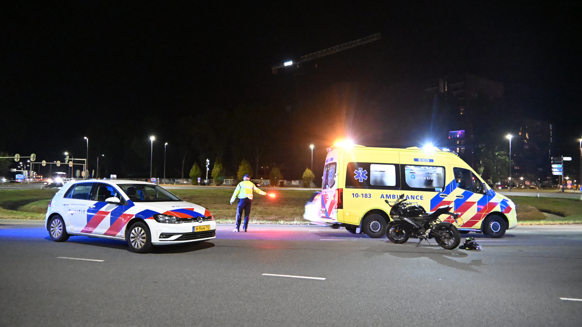 Politieauto en ambulance met zwaailichten bij nachtelijk ongeval; motorfiets en helm op de weg, agent regelt verkeer