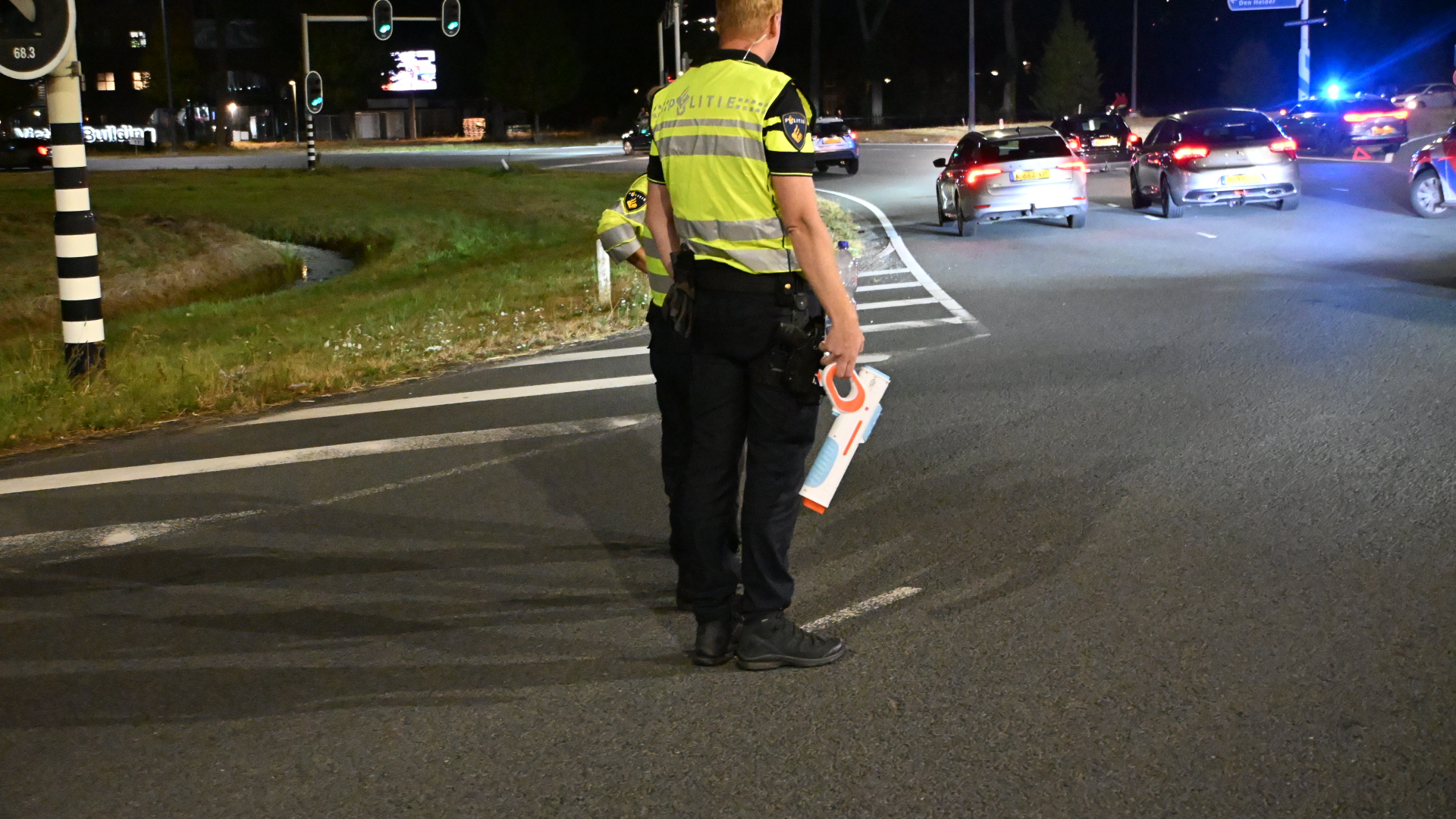 Politieagent in reflecterend vest regelt ’s avonds verkeer op rotonde, met auto’s en blauwe zwaailichten op de achtergrond
