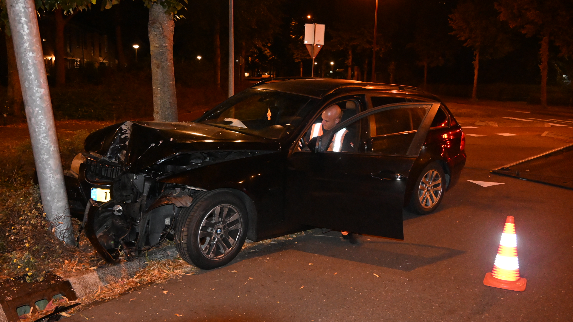 Zwarte auto met zware frontschade tegen een paal, ’s nachts; bestuurder stapt uit, met verkeerskegel op de weg