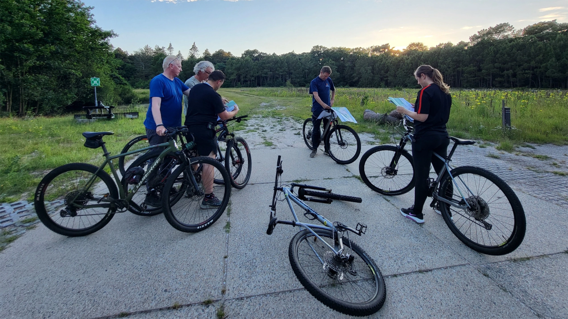 Groep mountainbikers pauzeert op een betonnen pad en bekijkt kaarten, met een fiets op de grond bij zonsondergang