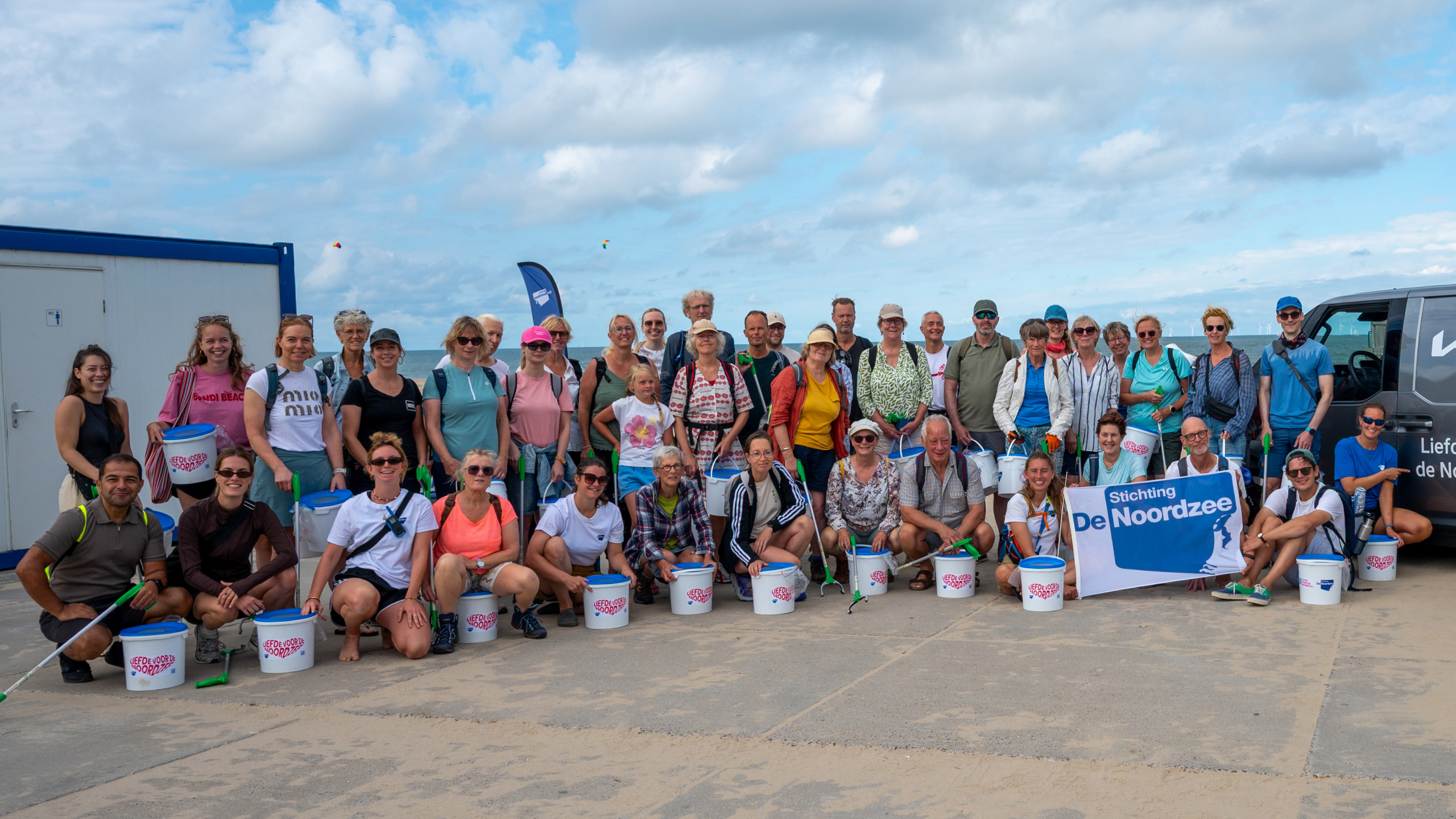 Groepsfoto van strandopruimers met emmers en grijpers; banner met tekst Stichting De Noordzee op de achtergrond