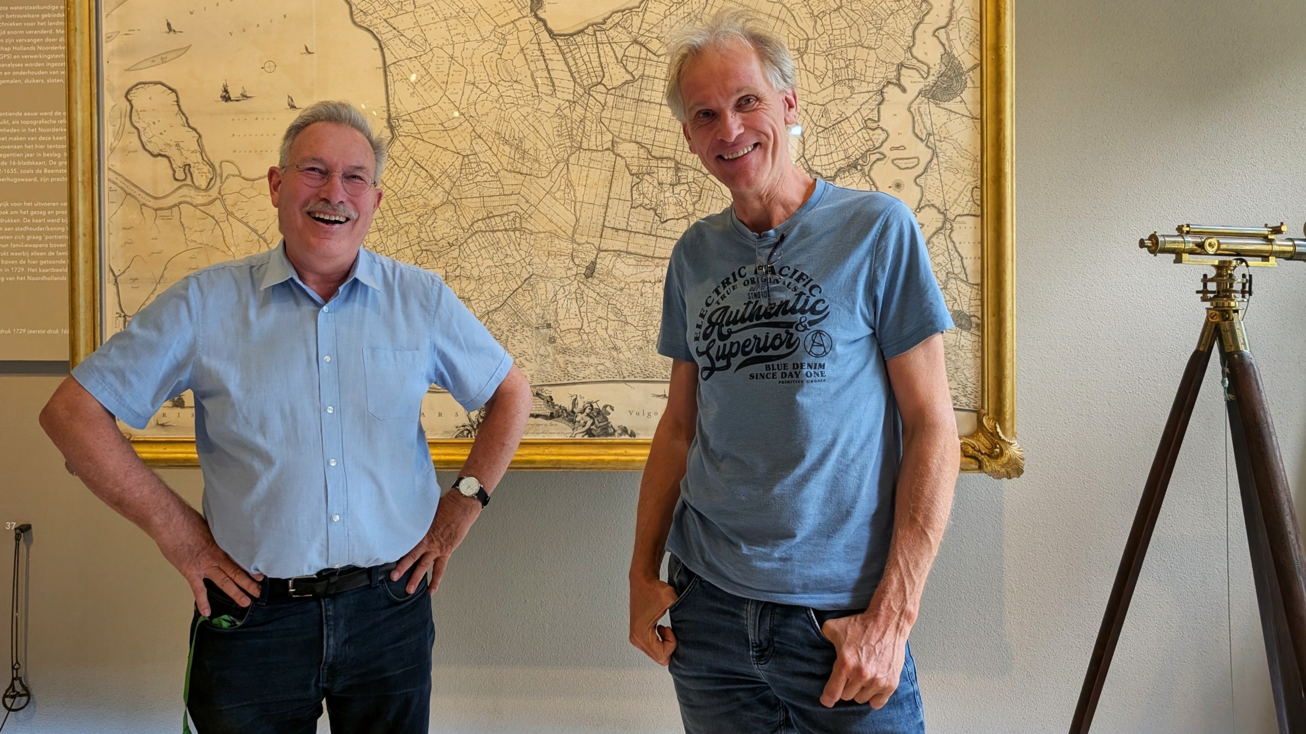 Twee lachende mannen poseren voor een grote ingelijste oude kaart, met rechts een landmeetkijker op statief