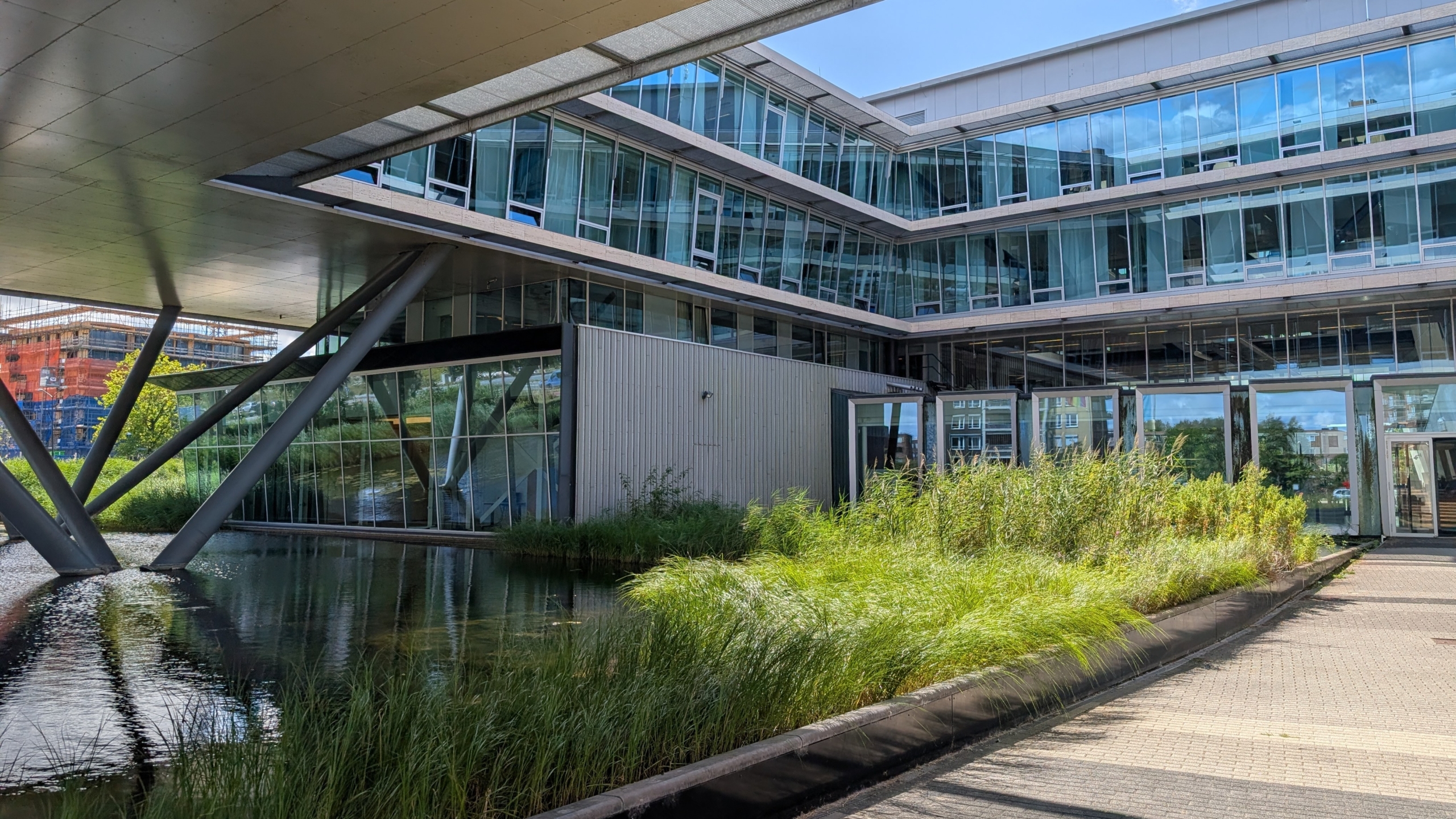 Modern kantoorgebouw met glazen gevels boven een vijver, stalen steunpilaren en riet langs een wandelpad