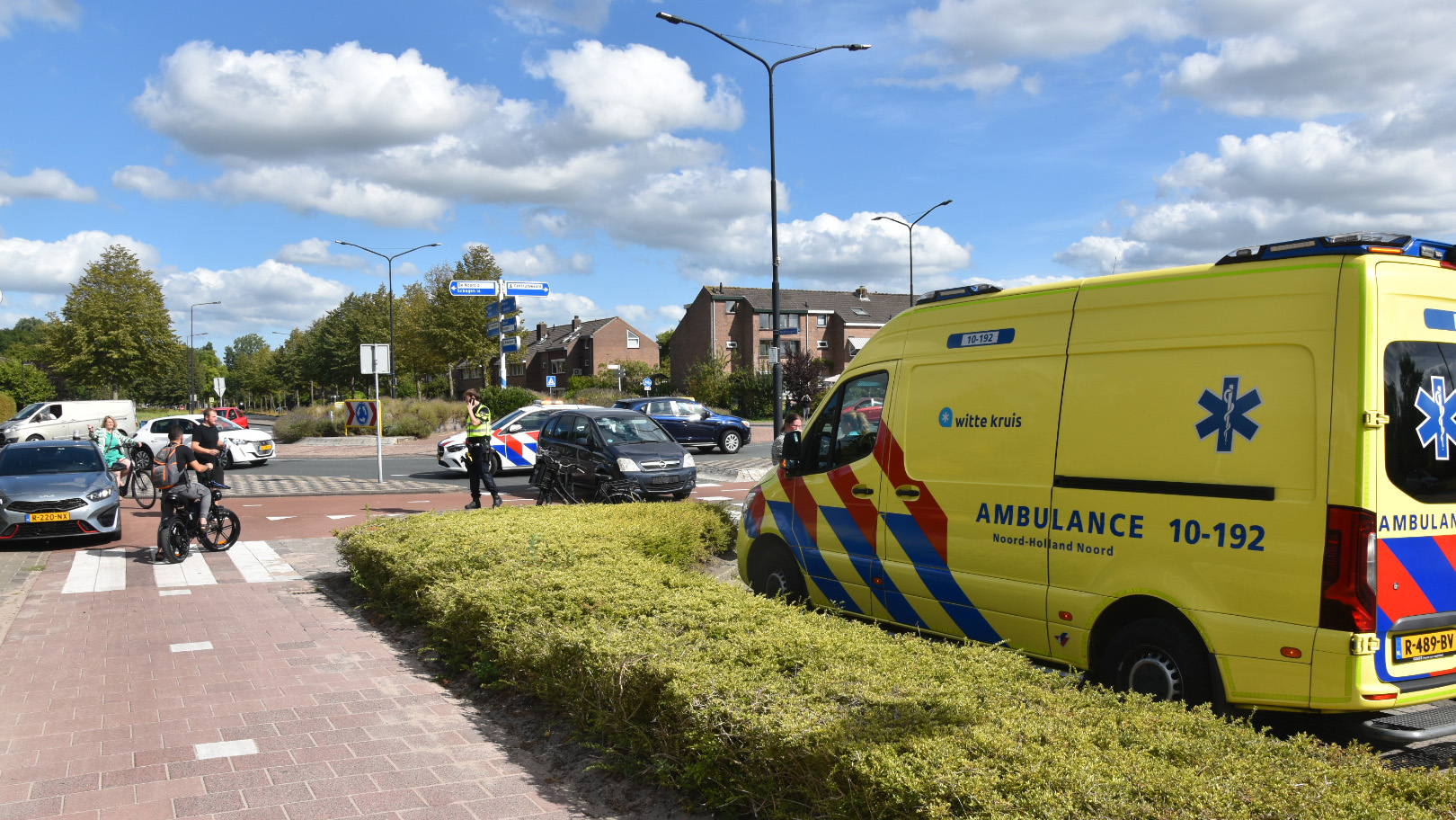 Gele ambulance en politieauto bij een rotonde, met omstanders en fietsers naast een beschadigde zwarte auto