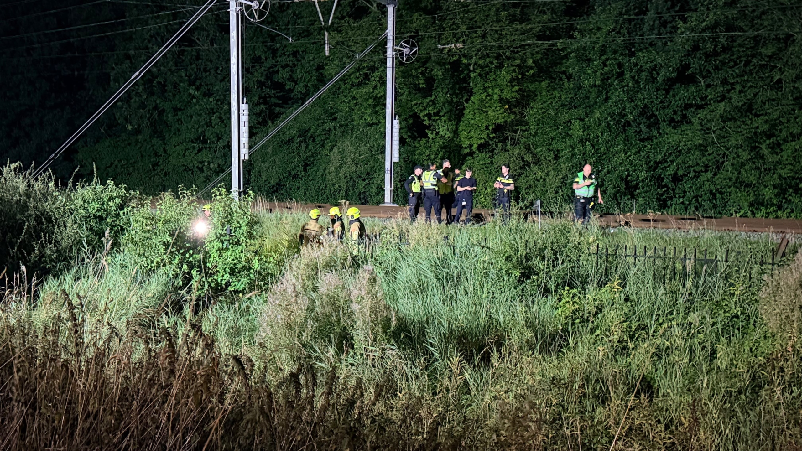 Hulpdiensten met zaklampen bij een spoorlijn langs dicht groen, vermoedelijk tijdens een nachtelijke zoekactie