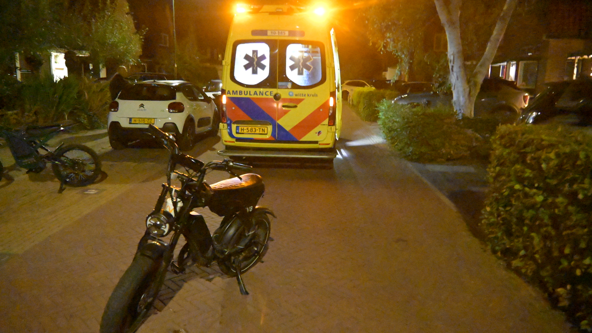 Ambulance met zwaailichten staat ’s avonds in woonstraat; brommer op voorgrond en fiets ligt op de grond links