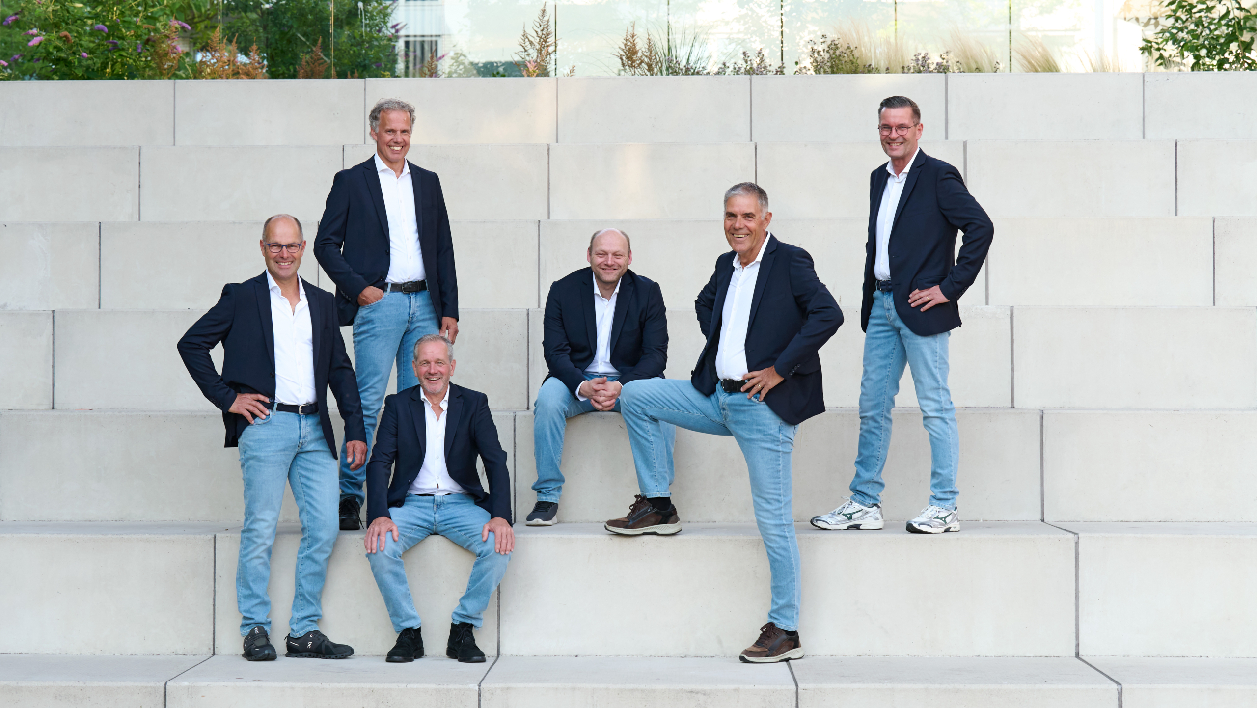 Zes mannen in colbert en spijkerbroek poseren op betonnen traptreden in een moderne buitenruimte