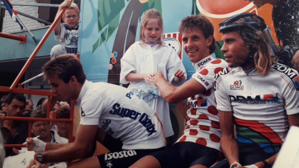 Wielrenners in wedstrijdshirts op een podiumwagen, omringd door publiek; een meisje staat ertussen en kijkt toe