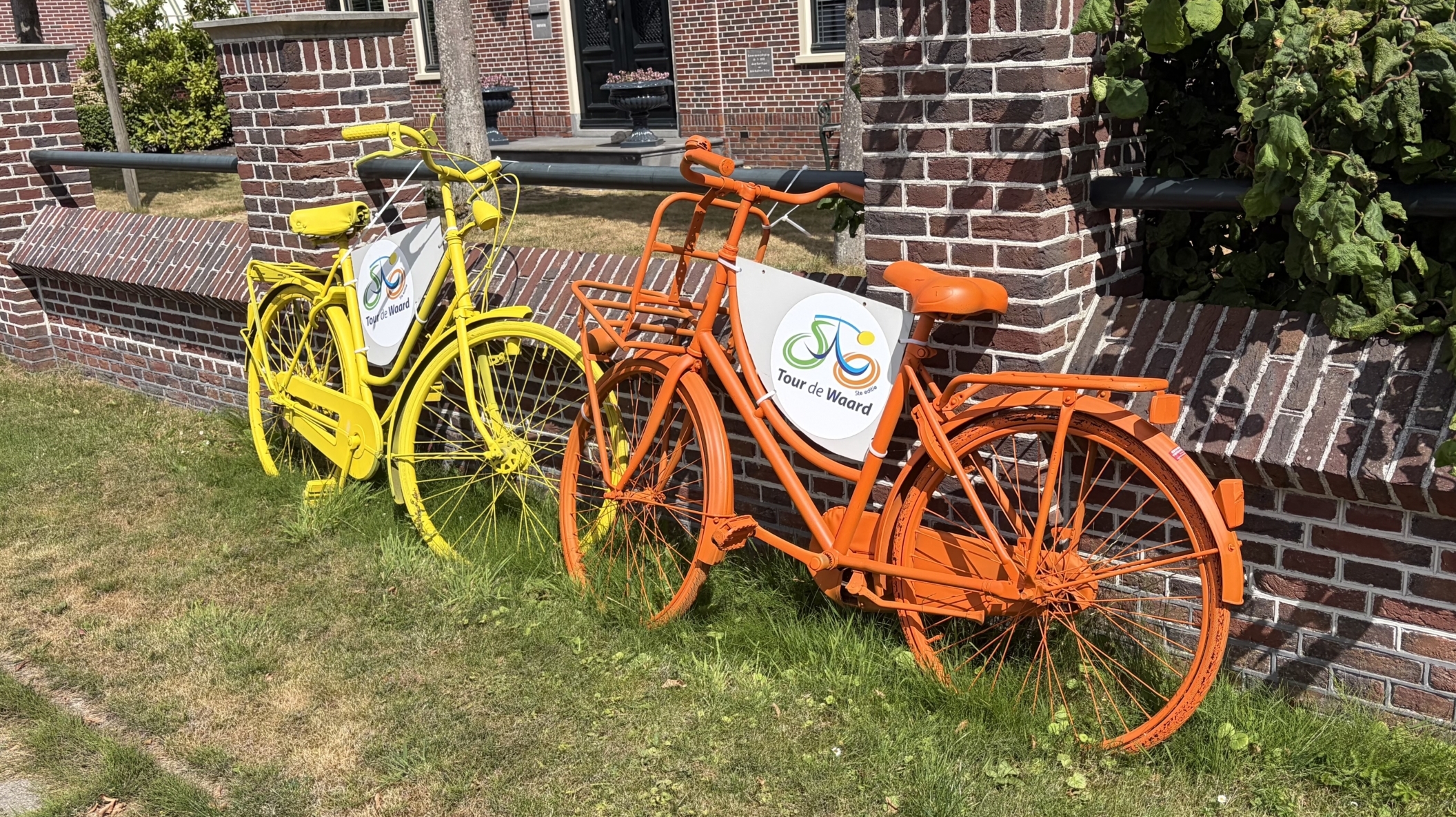 Gele en oranje fiets met bordje “Tour de Waard” staan tegen een bakstenen muur in het gras