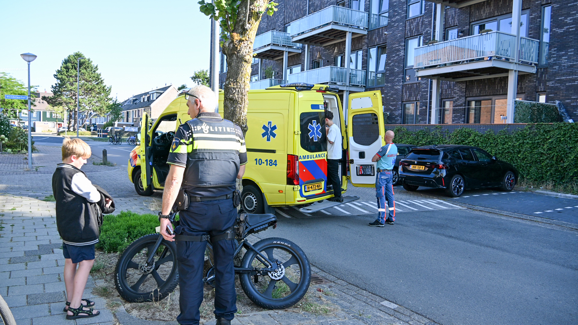 Politieagent op fiets bij gele ambulance met open achterdeuren, terwijl een ambulancebroeder helpt op straat