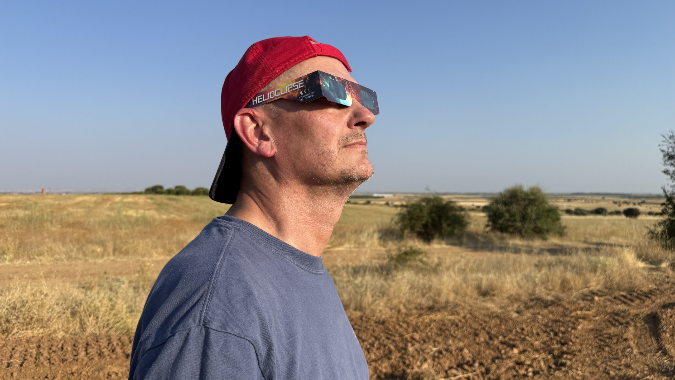 Man met rode pet kijkt omhoog met eclipsbril in droog grasland onder heldere blauwe lucht
