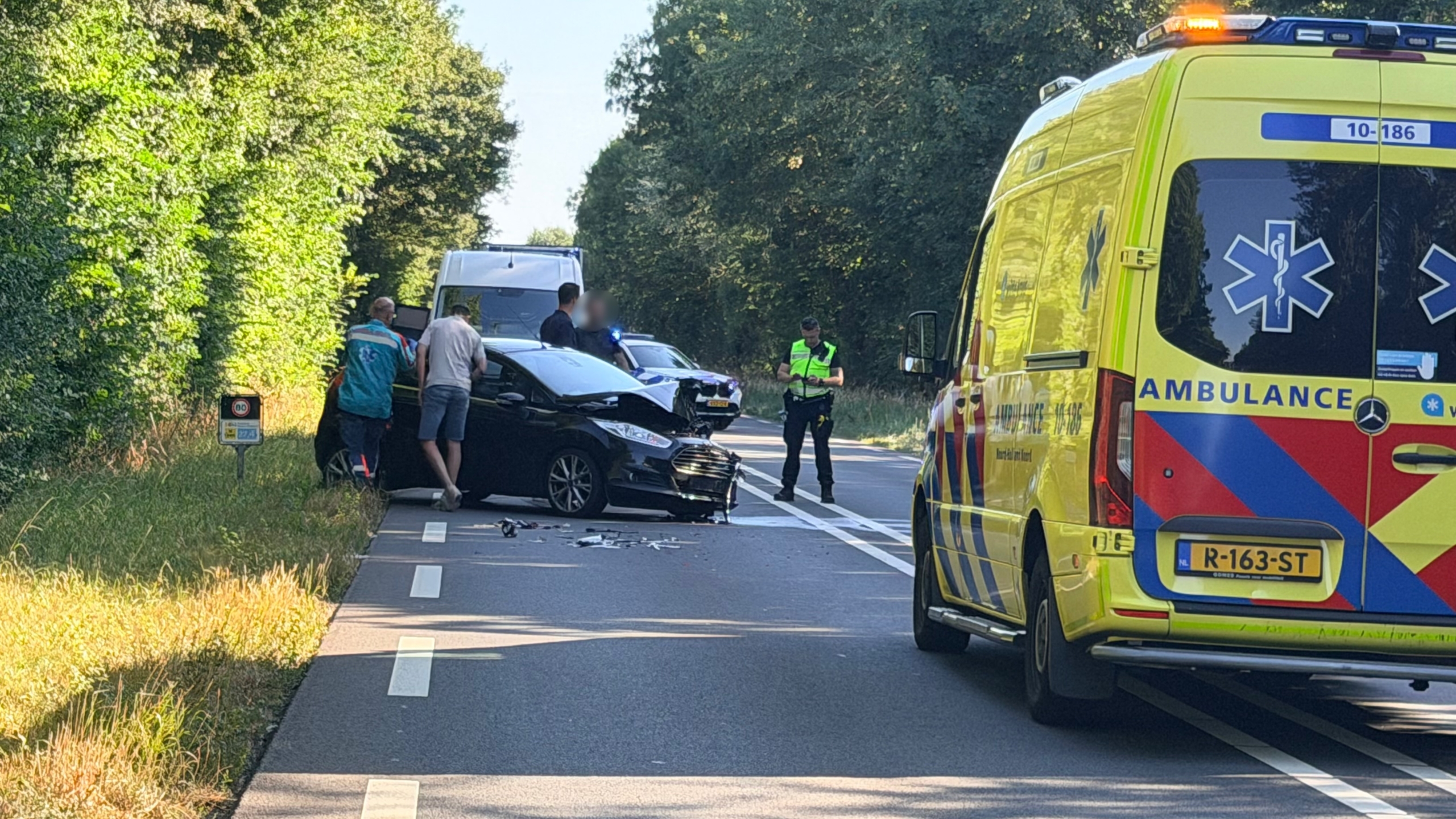 Ambulance en politie bij auto-ongeluk op bosrijke weg, met beschadigde zwarte auto en brokstukken op het asfalt