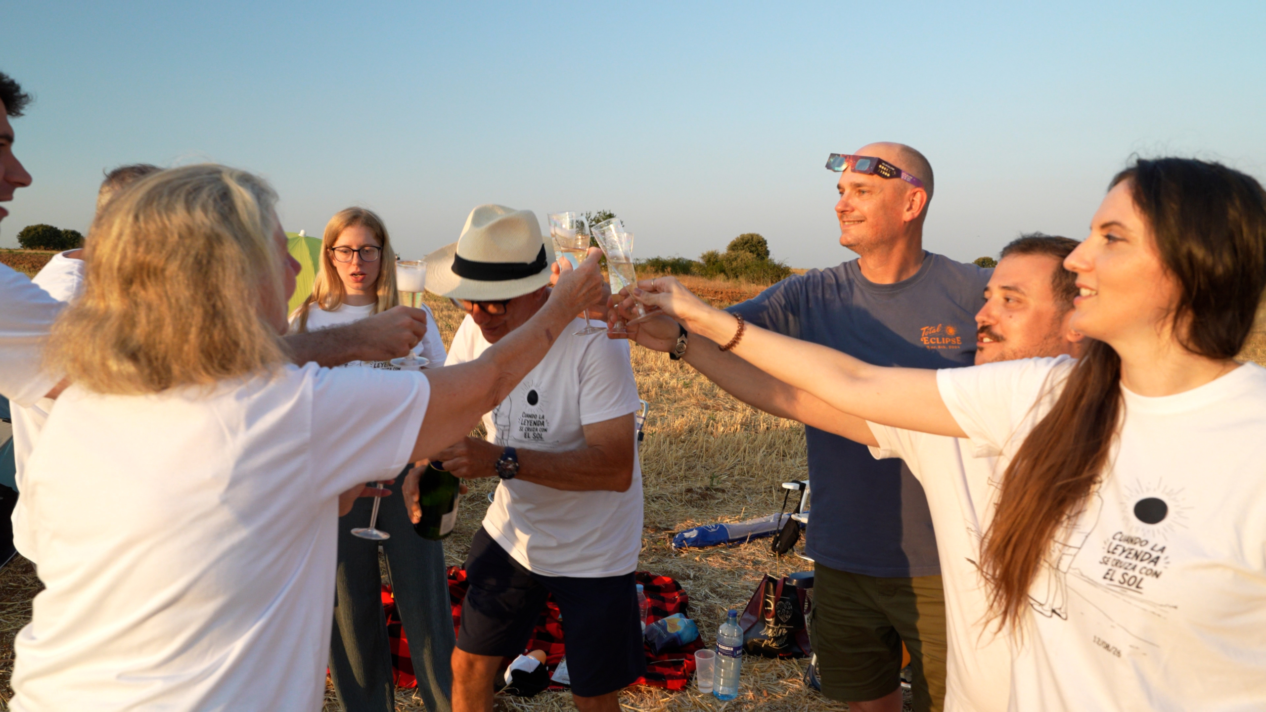 Groep mensen proost met champagneglazen op een veld bij zonsondergang, een man draagt eclipsbril op zijn hoofd