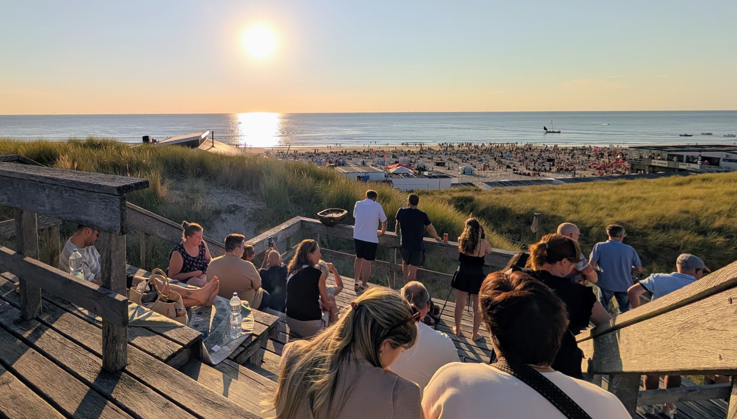 Mensen zitten op houten trappen in de duinen en kijken naar een druk strand en de zee bij zonsondergang