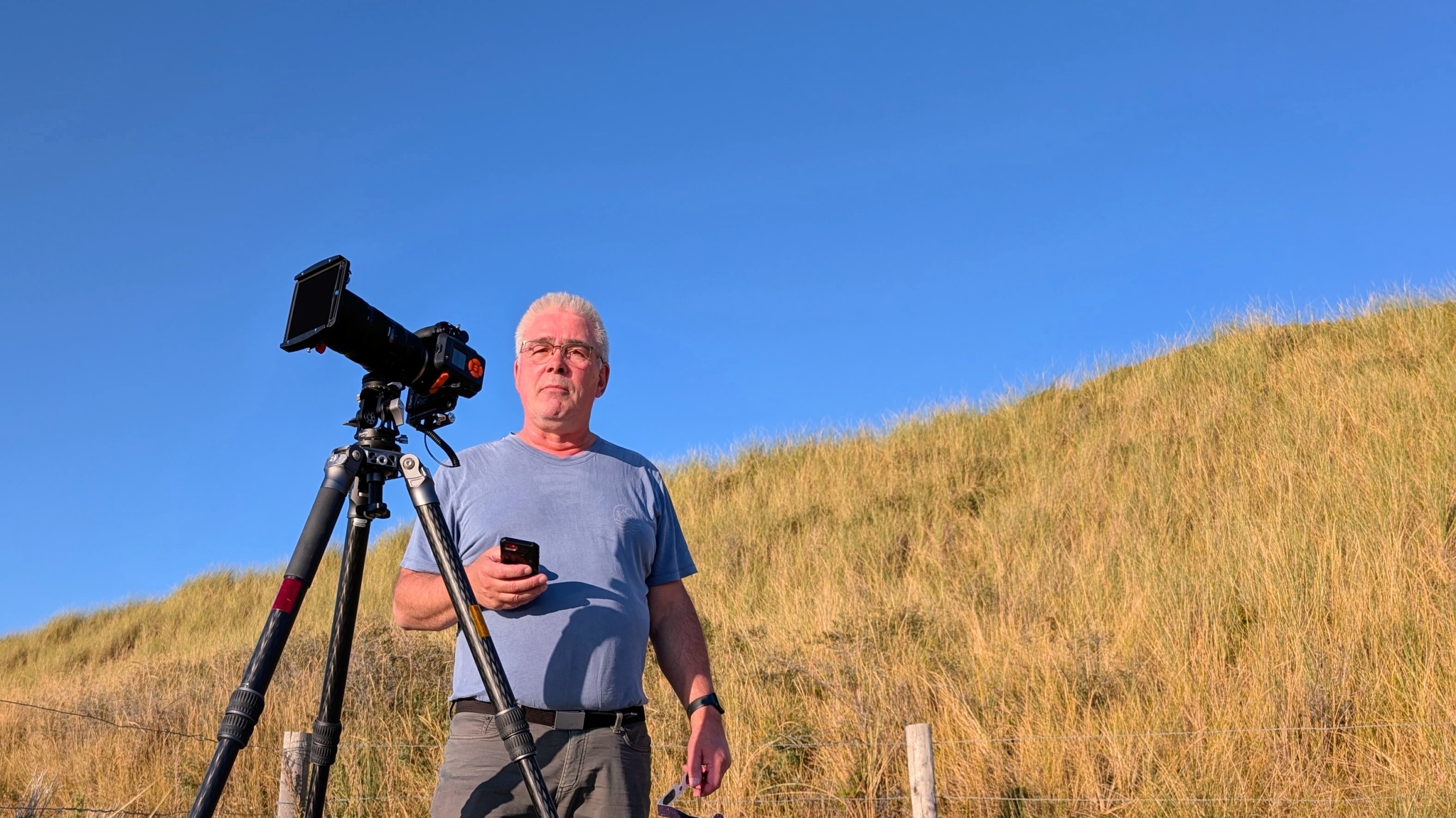 Man met smartphone naast camera op statief in duinlandschap met hoog gras onder een heldere blauwe lucht