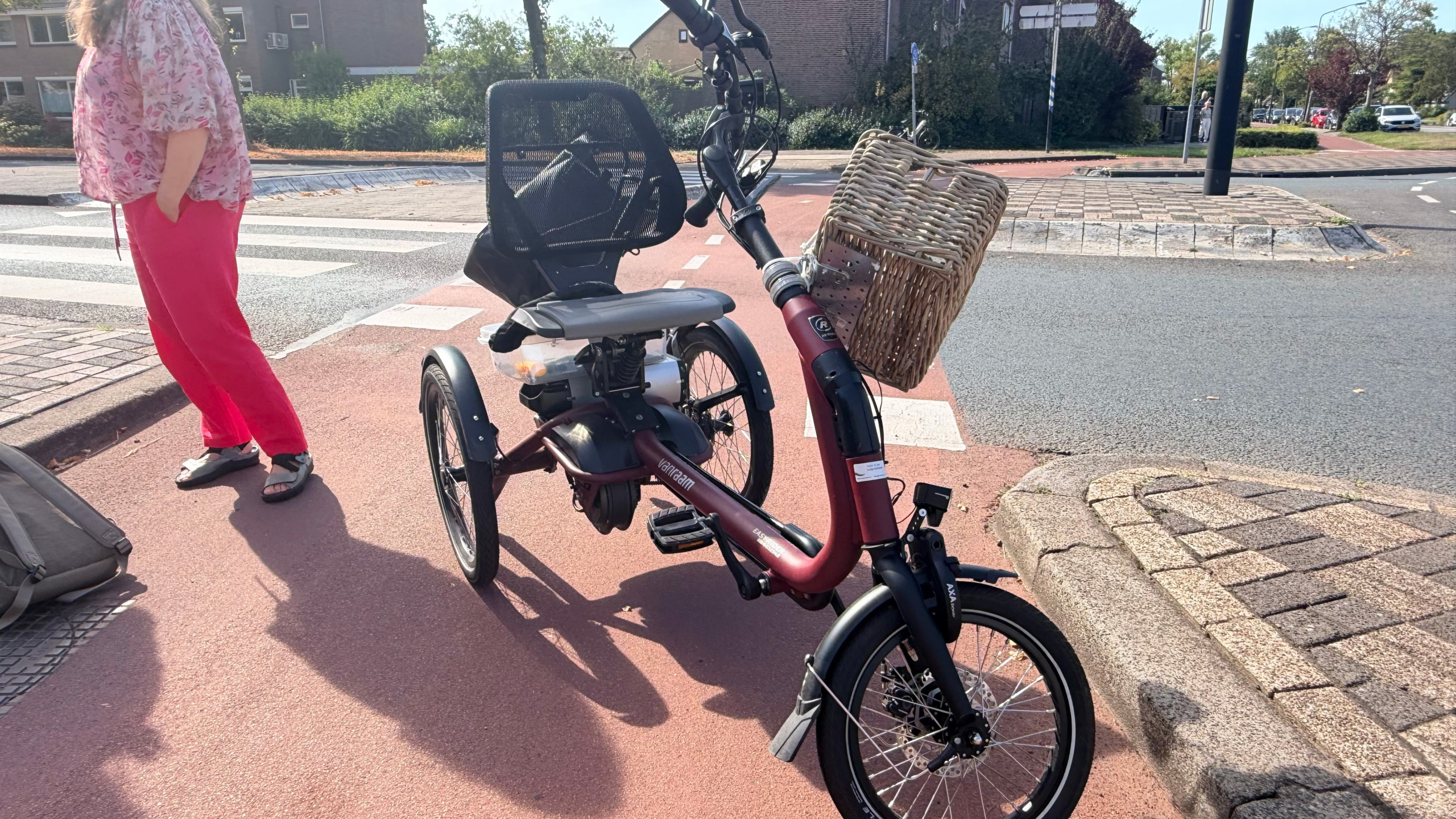 Driewieler met lage instap, mandje voorop en rugleuning, geparkeerd op een fietspad bij een kruising