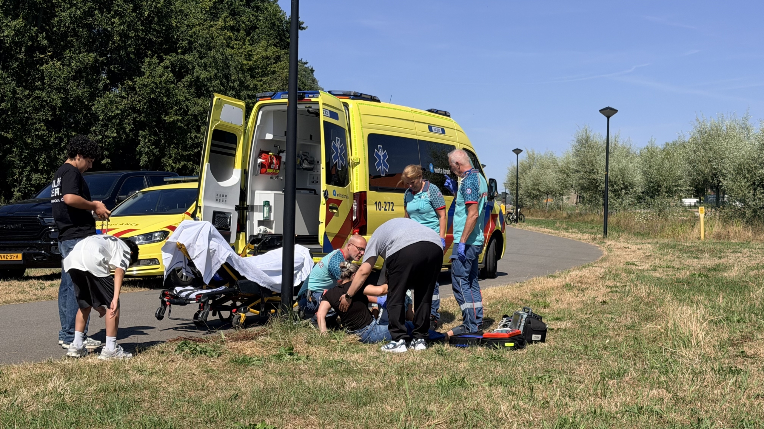 Ambulance met open achterdeuren; hulpverleners behandelen iemand op de grond naast een brancard langs een fietspad